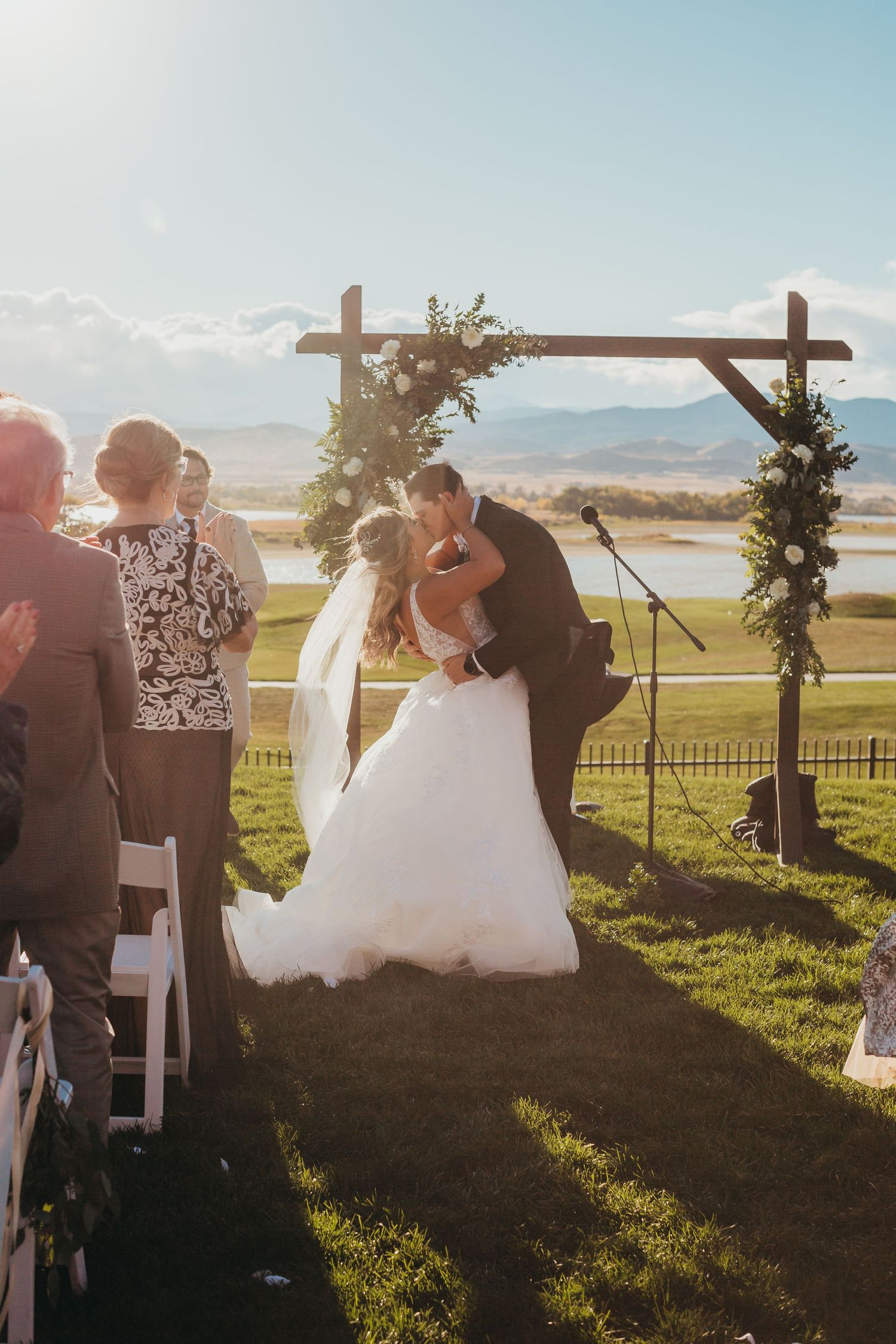 Romantic outdoor wedding ceremony under a wooden arch adorned with greenery at sunset in a scenic mountain setting.