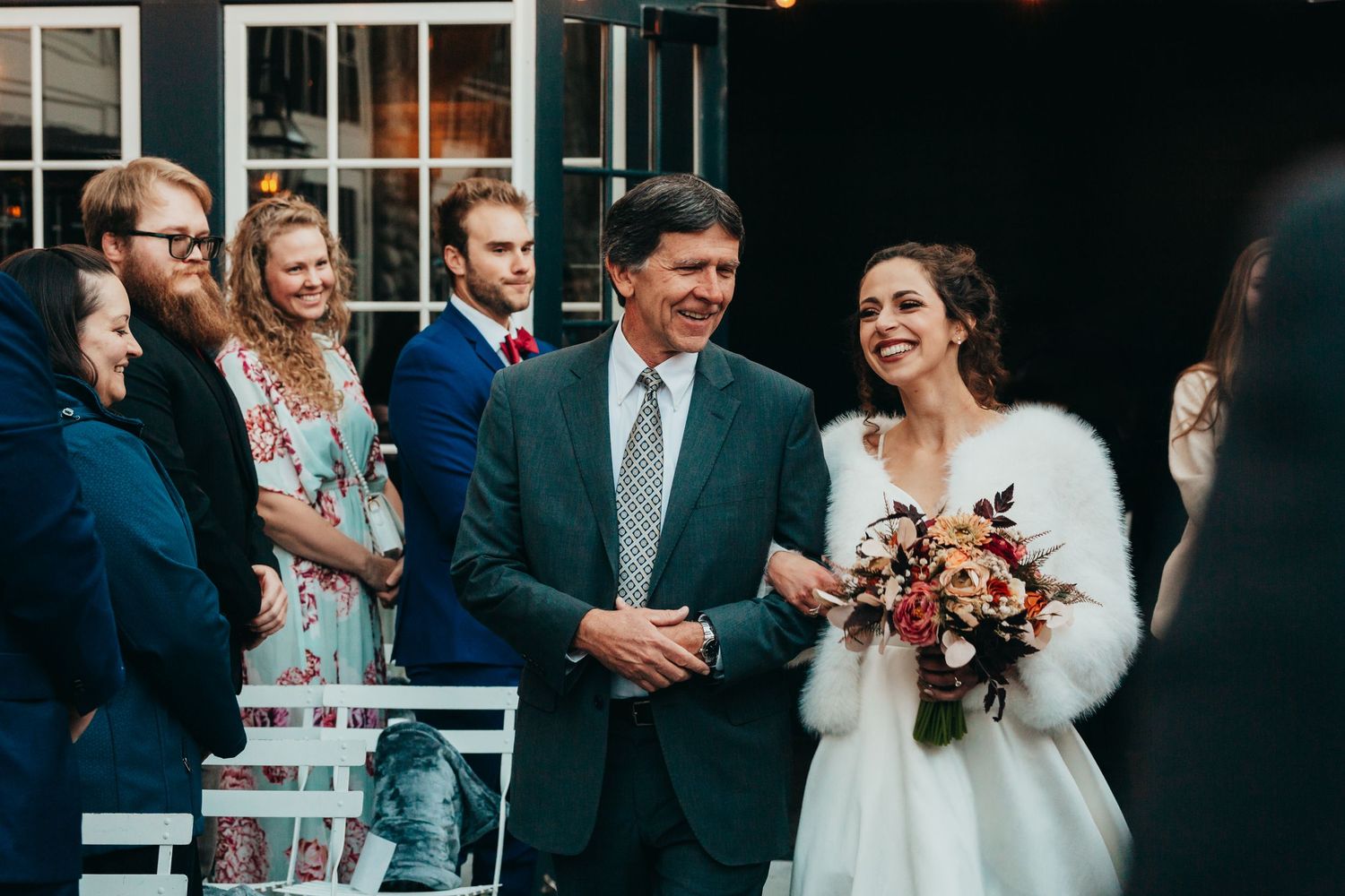 Joyful wedding party gathering outdoors with the bride wearing a white fur wrap and holding a bouquet.