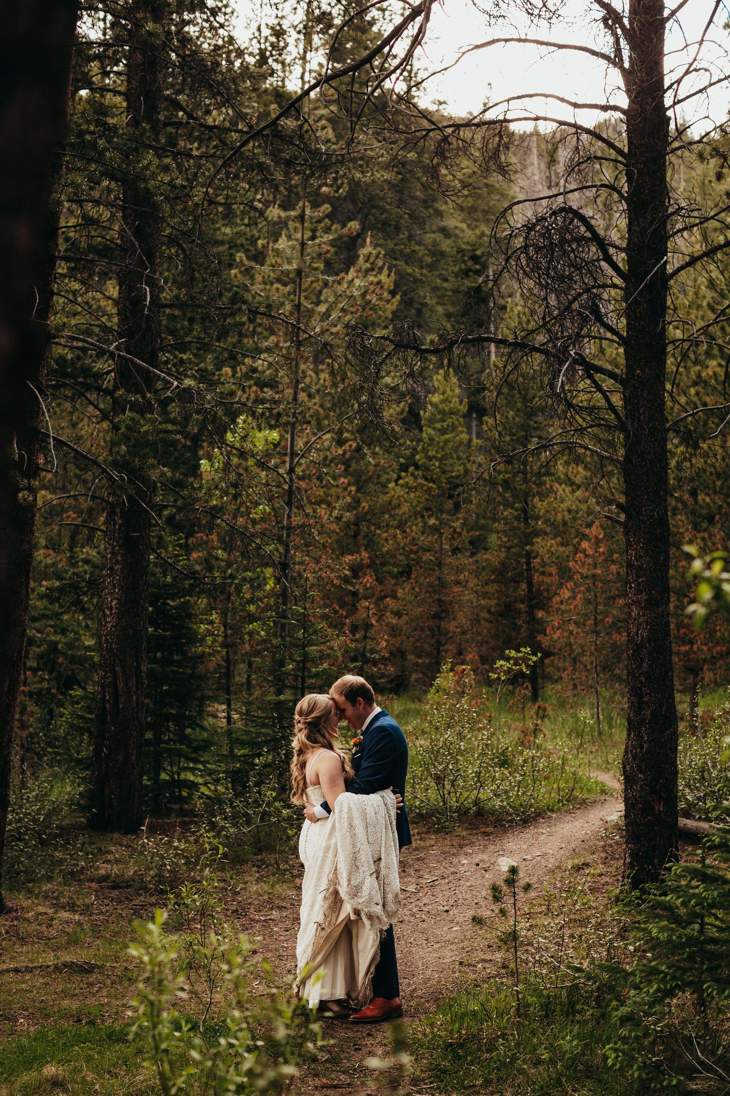 A couple shares an intimate moment on a forest path surrounded by tall evergreen trees.