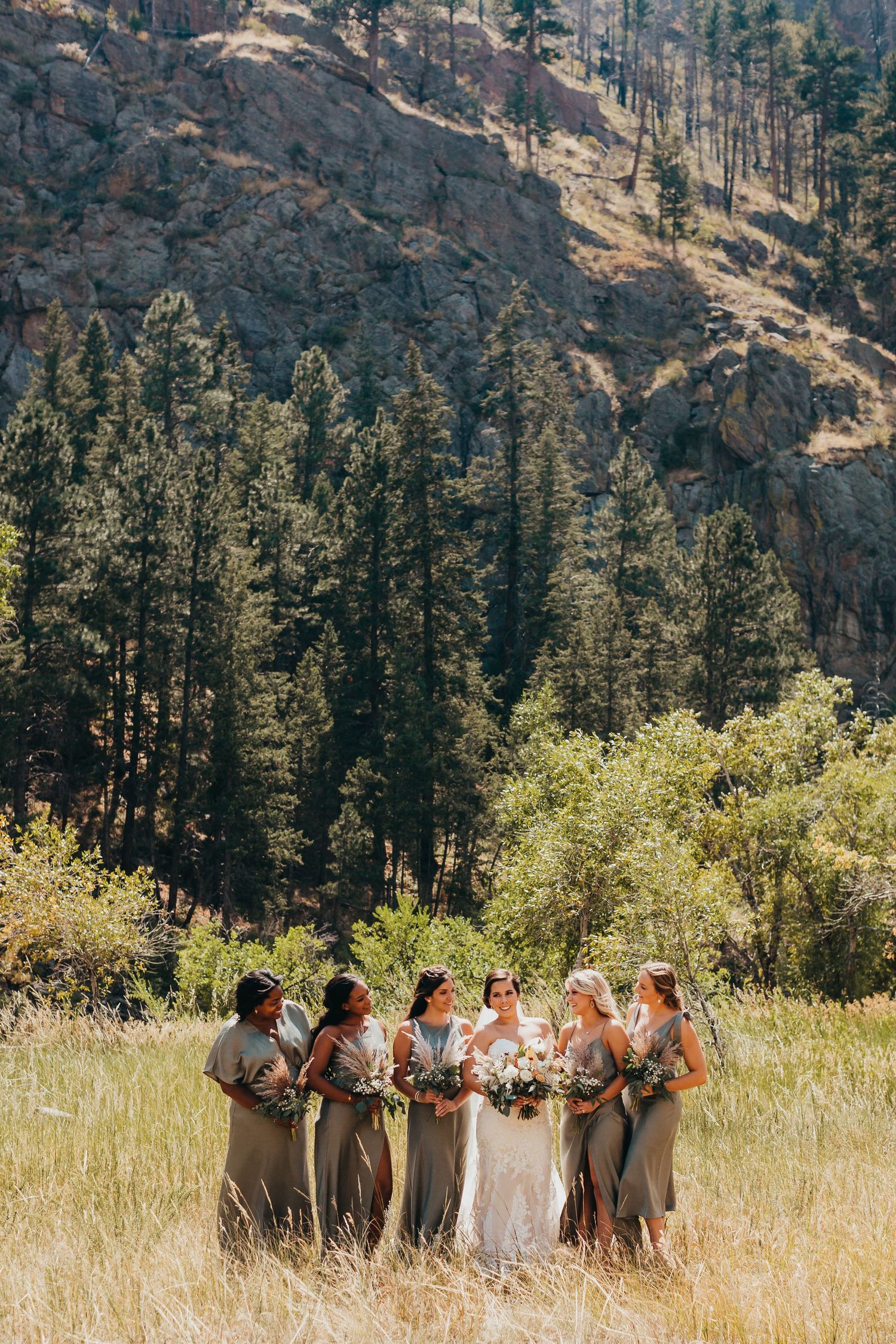 Bridesmaids and bride pose together in golden field with mountain landscape and evergreen trees.