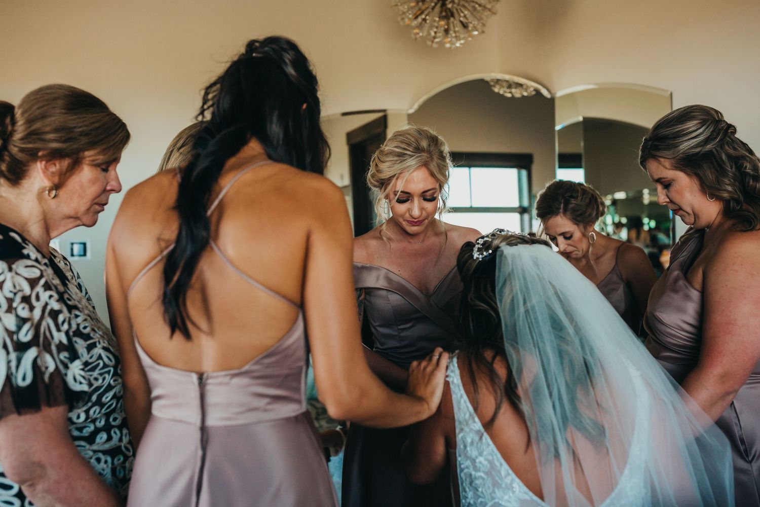 Bridesmaids in mauve dresses gather around the bride helping with final wedding preparations.