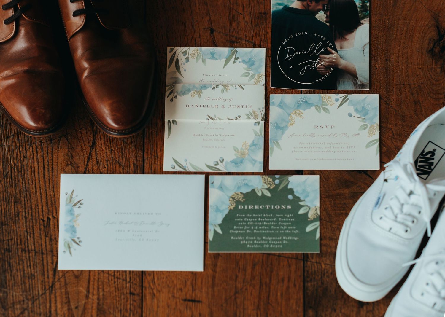 Wedding invitation suite with brown shoes and white sneakers on wooden floor.