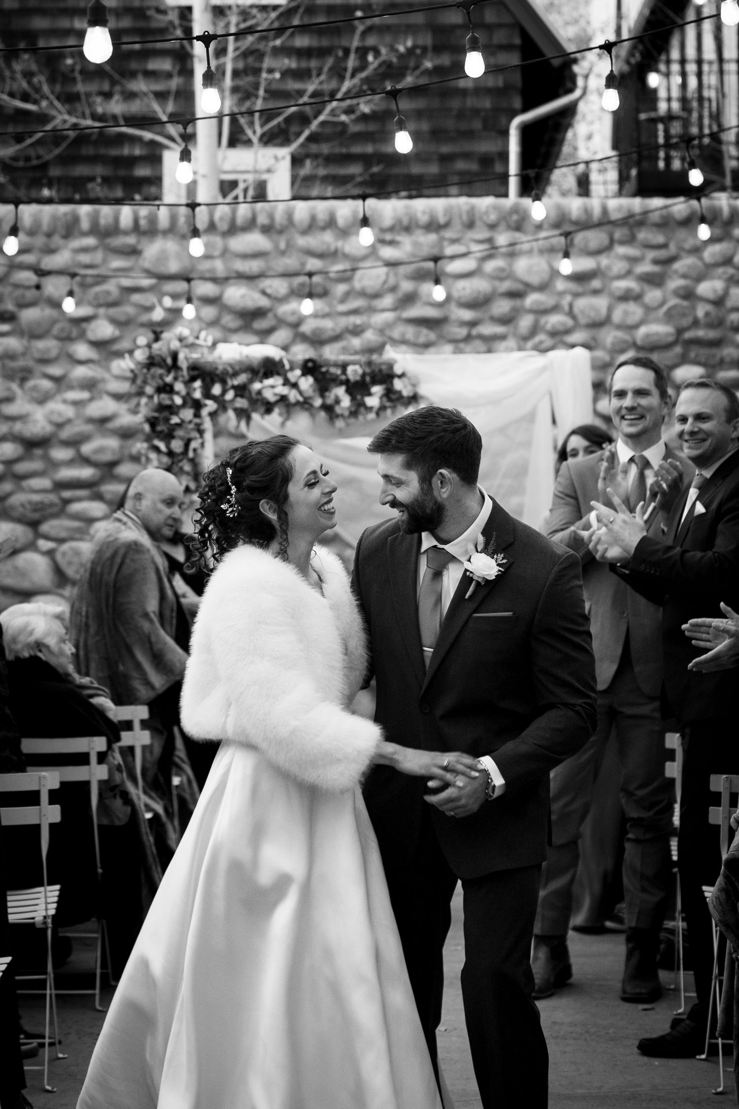 A joyful wedding moment against a stone wall backdrop with string lights and floral decor.