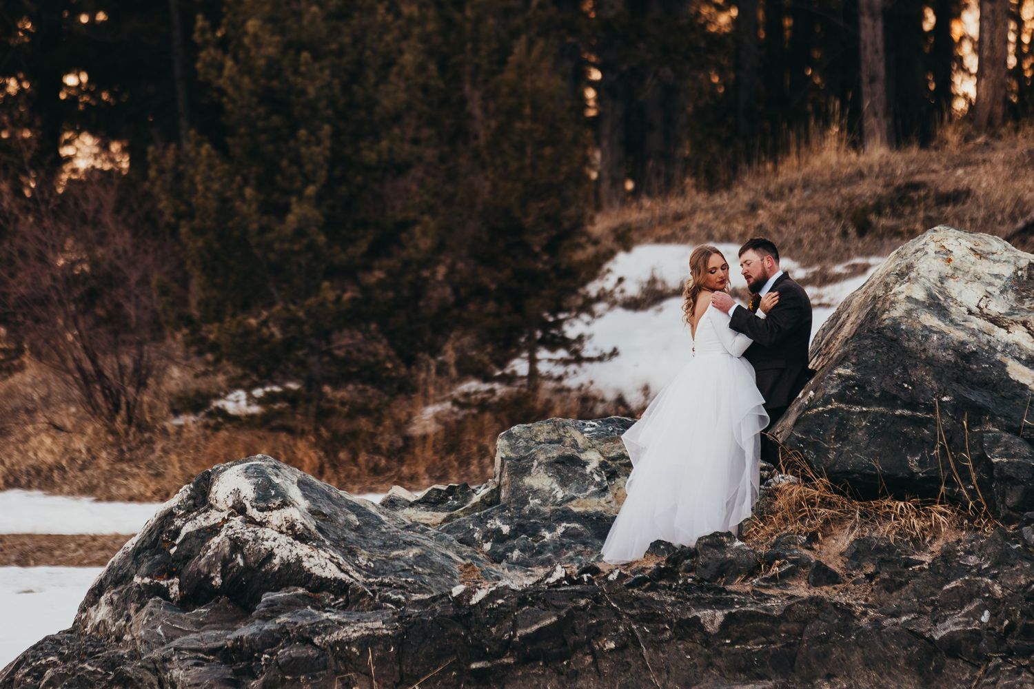 A wedding couple posing on rocky terrain with snow and forest in the background.