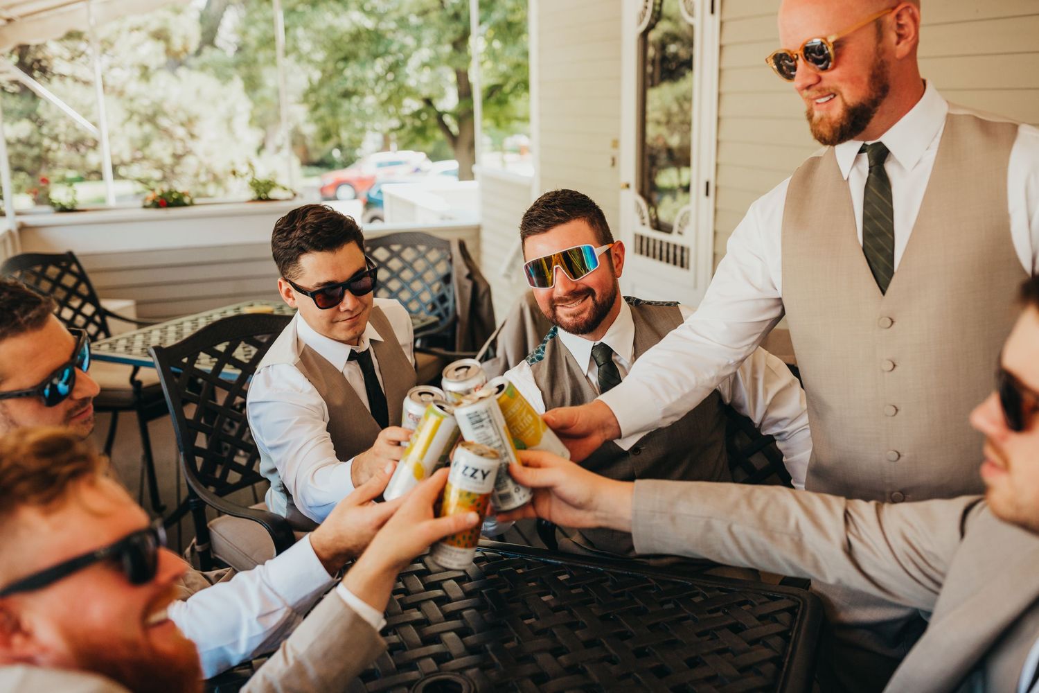 Groomsmen in sunglasses toasting with drinks on a porch during wedding celebrations.