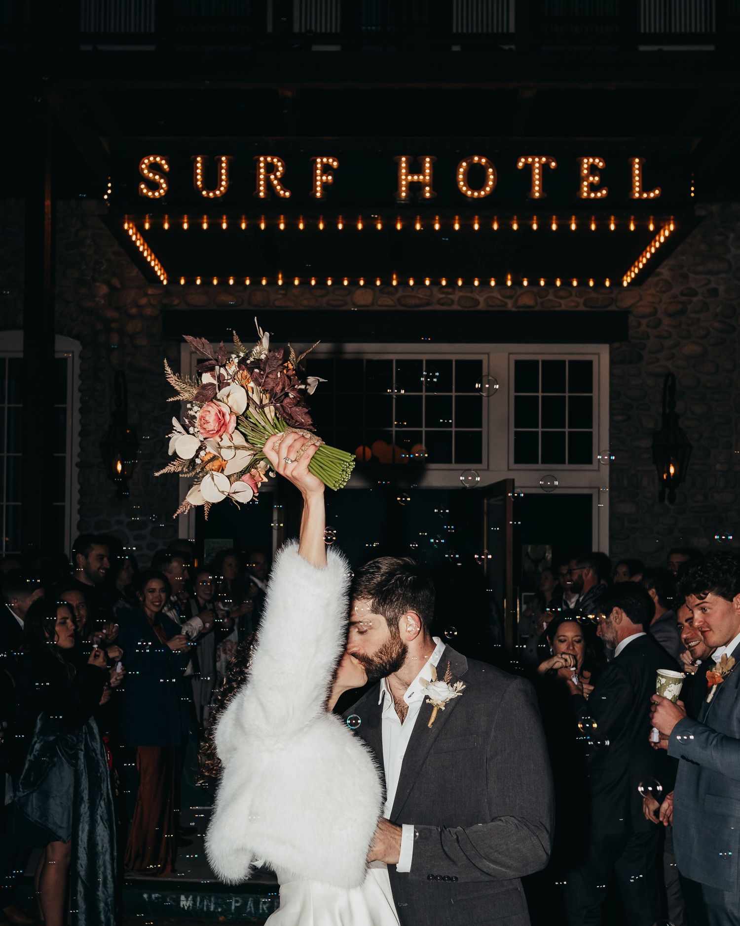 Couple with bouquet sharing a moment under the illuminated Surf Hotel sign at night.