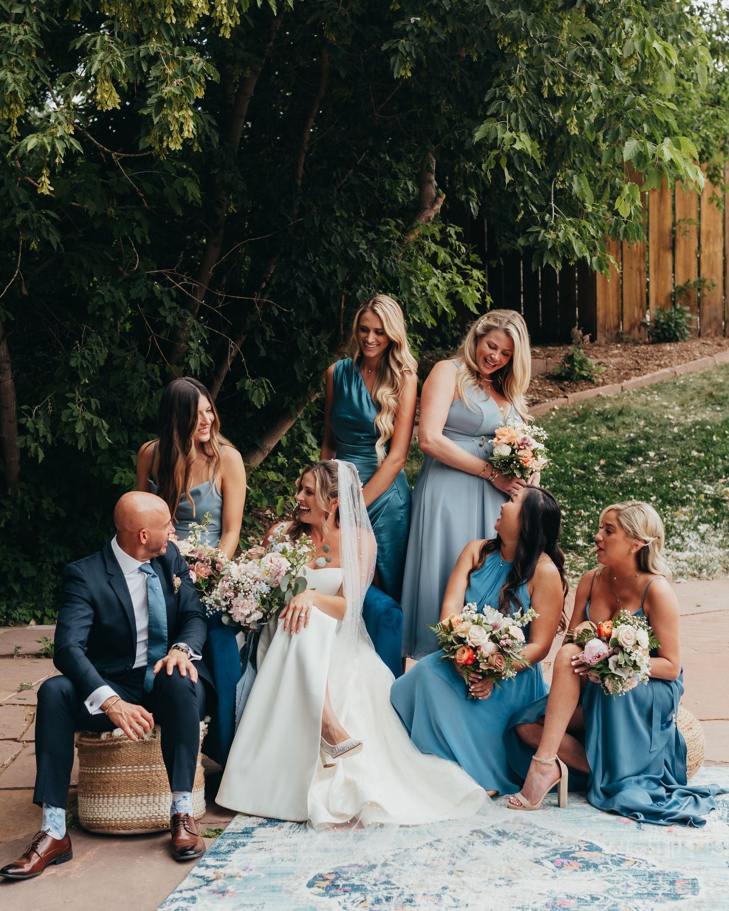 Wedding party relaxes on patio furniture in coordinated blue attire while holding bouquets.