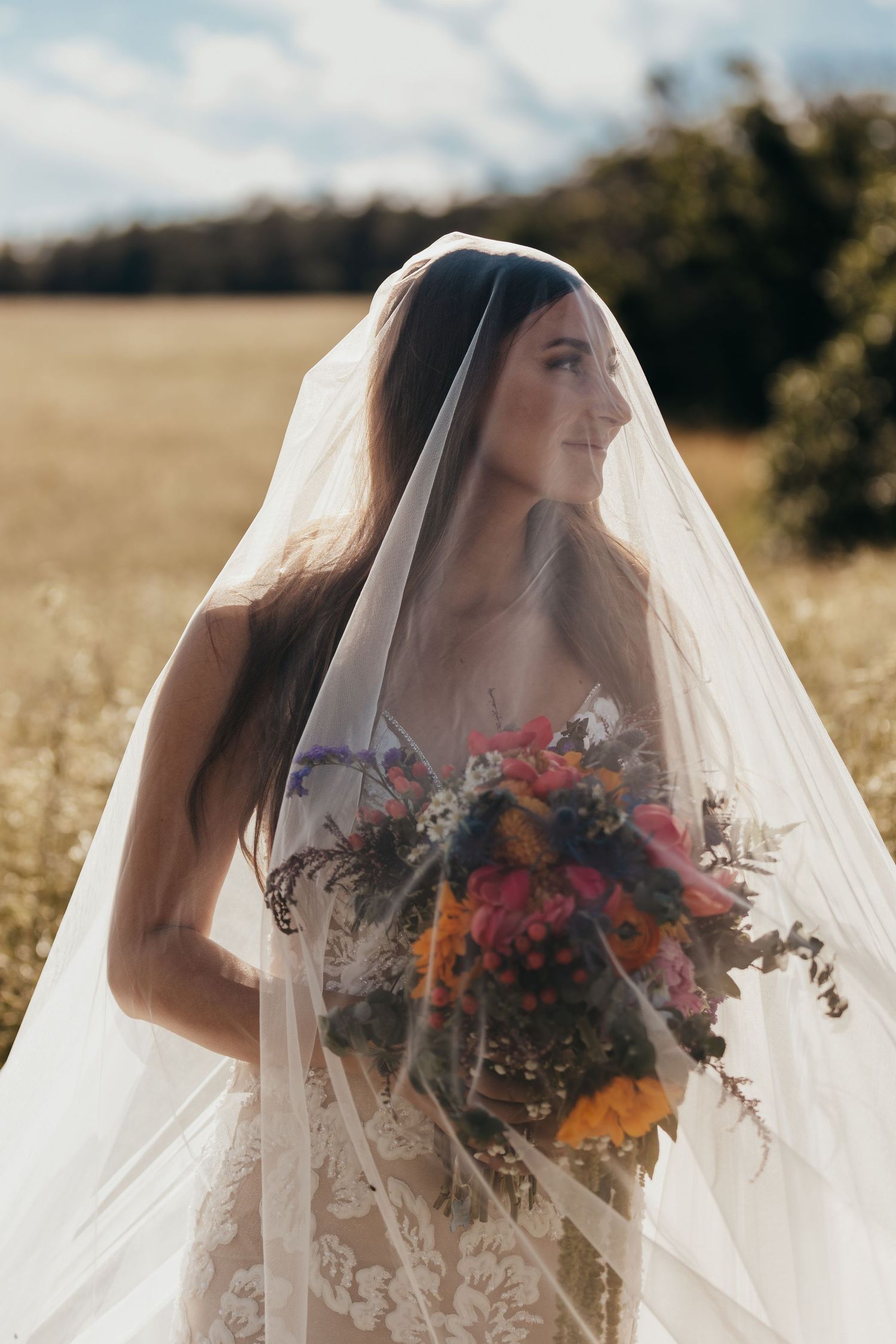 Bride in lace gown holds vibrant autumn bouquet while standing in golden field.