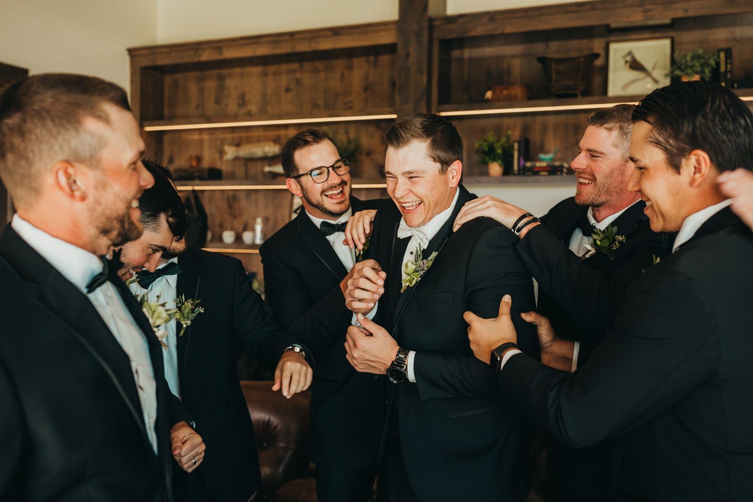 A group of groomsmen in suits laughing and sharing drinks at a wedding celebration.