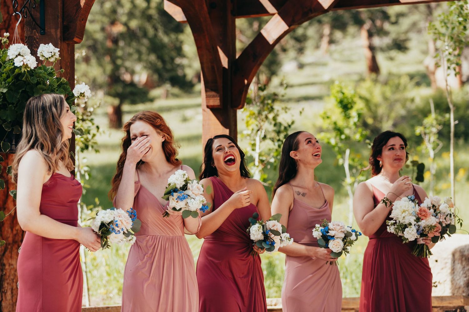 Bridesmaids in pink dresses share joyful laughter while holding bouquets during outdoor wedding.