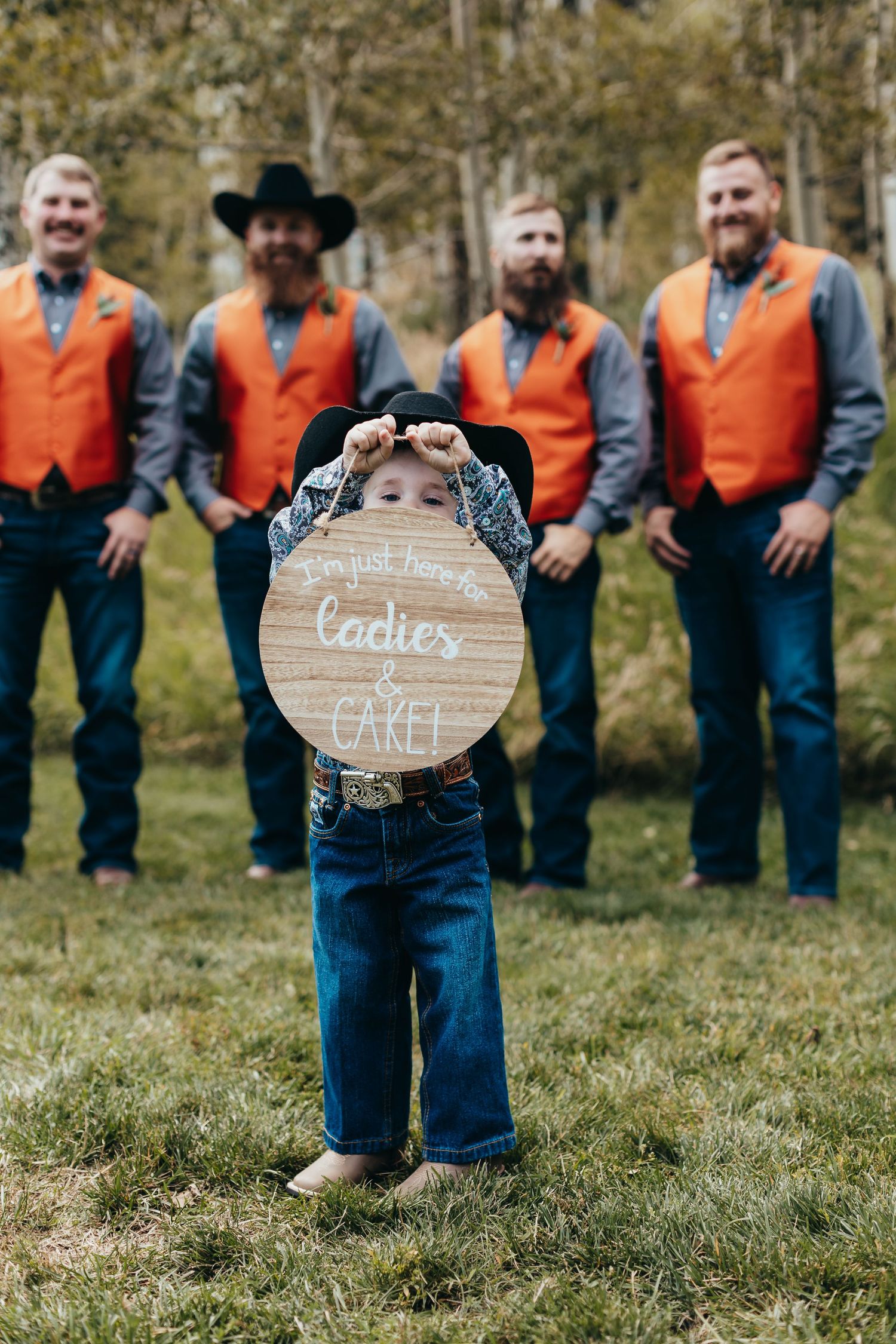 A wedding party photo with groomsmen in orange vests behind a rustic wooden sign.