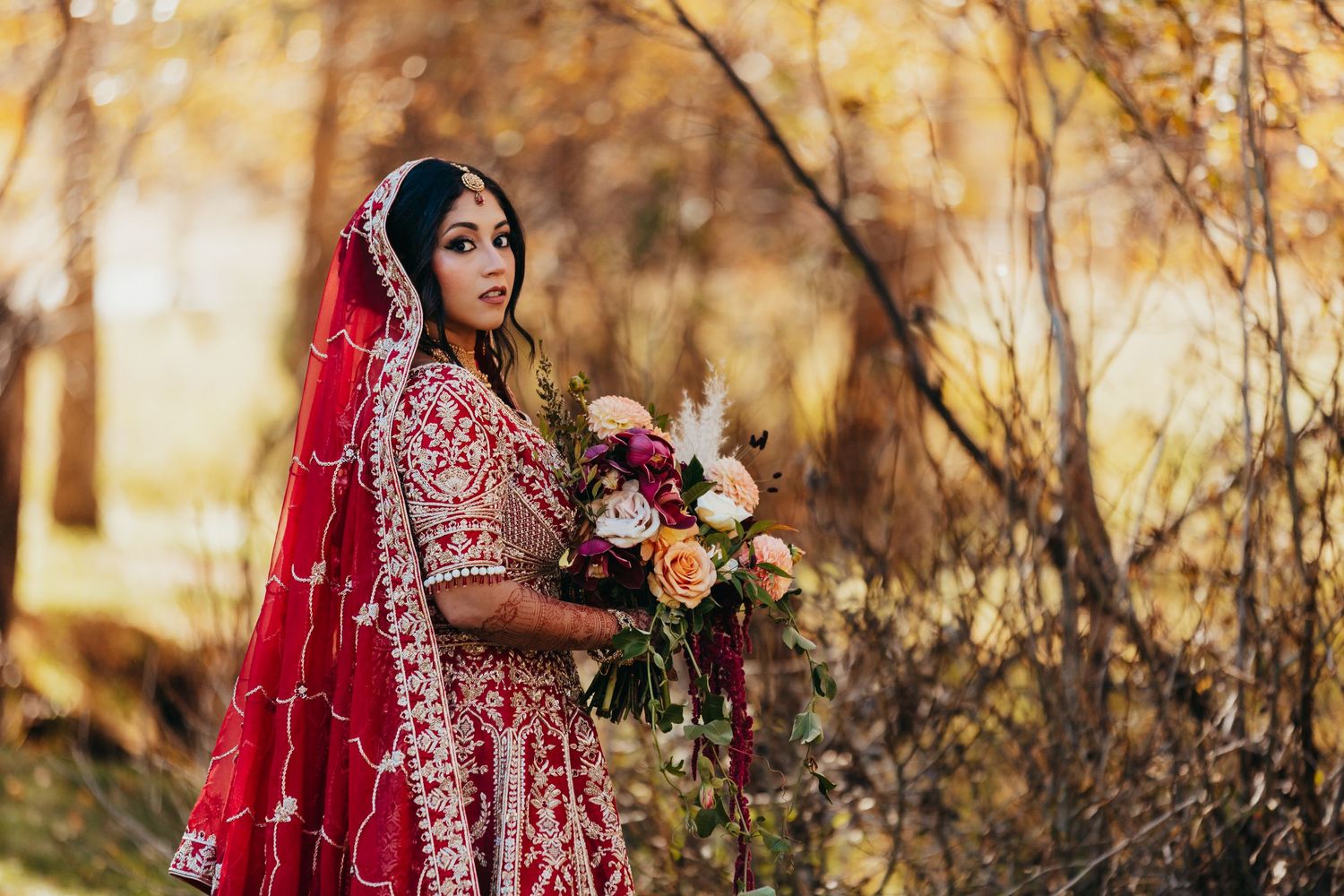 Autumn wedding portrait featuring traditional red bridal attire and colorful floral bouquet against golden foliage.