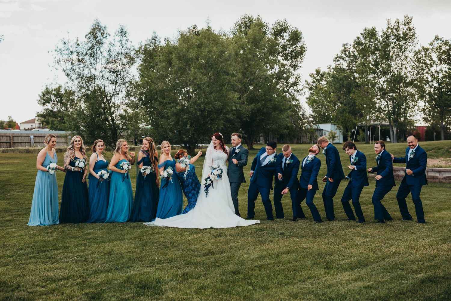 Wedding party poses in a fun line on grass with bridesmaids in blue and groomsmen in navy suits.