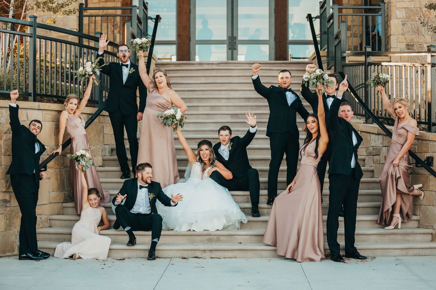 Wedding party strikes playful poses on stone steps in formal attire.