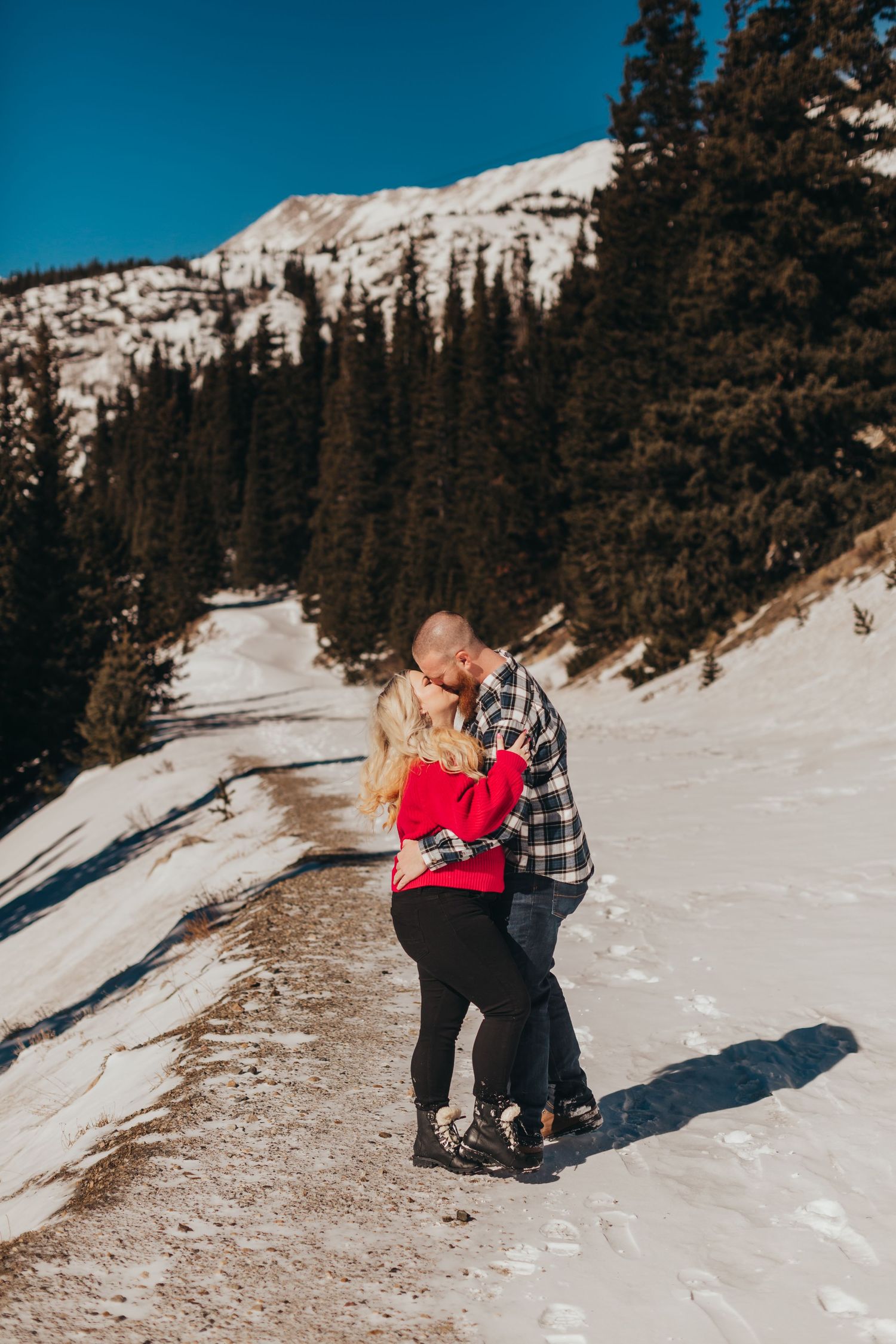 Couple shares romantic moment on snowy mountain trail with evergreen forest backdrop.