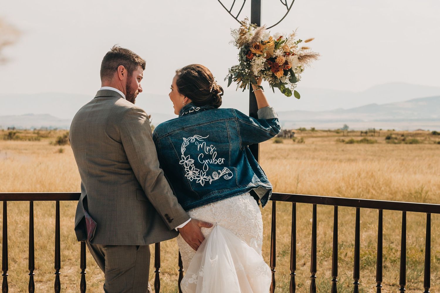 Wedding couple embraces while wearing a custom denim jacket in a rustic outdoor setting.