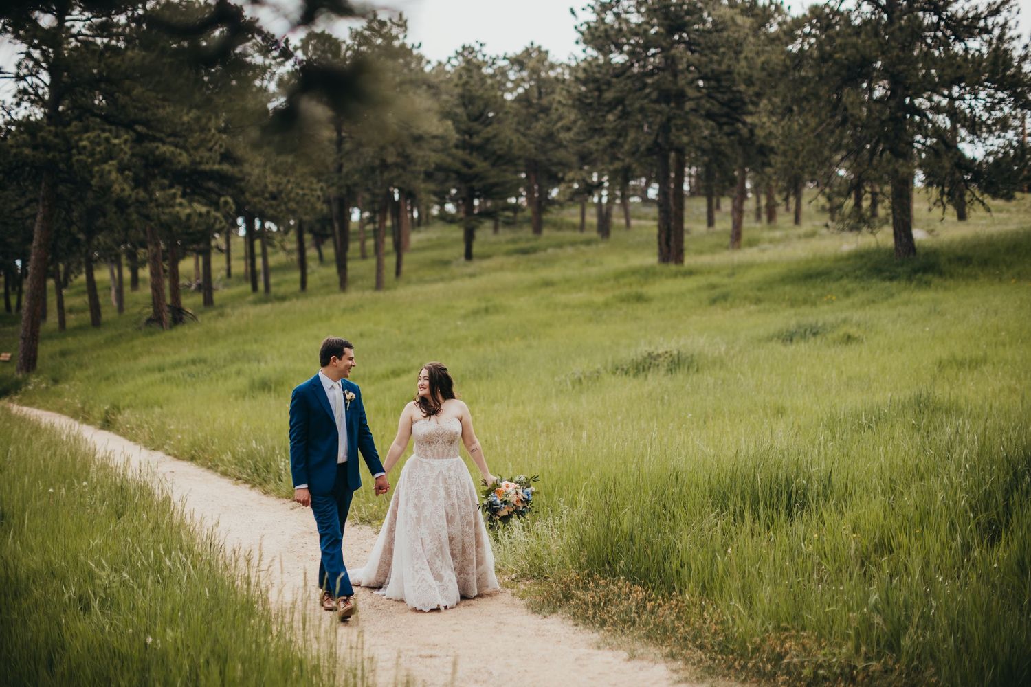 Romantic wedding portrait of couple walking down a mountain path surrounded by tall grass.