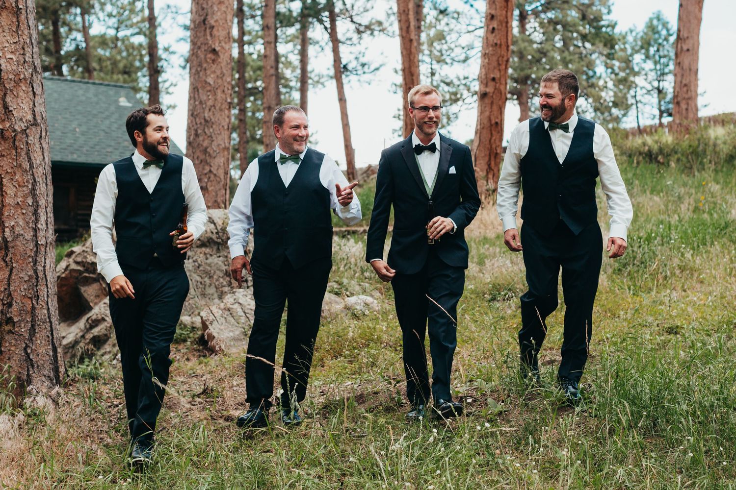 Groomsmen in matching black vests and bowties walking through pine trees.