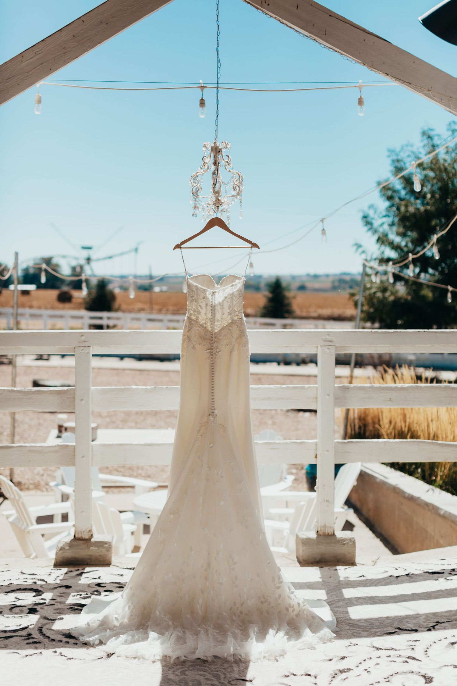 White lace wedding dress hangs from ornate chandelier against blue sky outdoor setting.