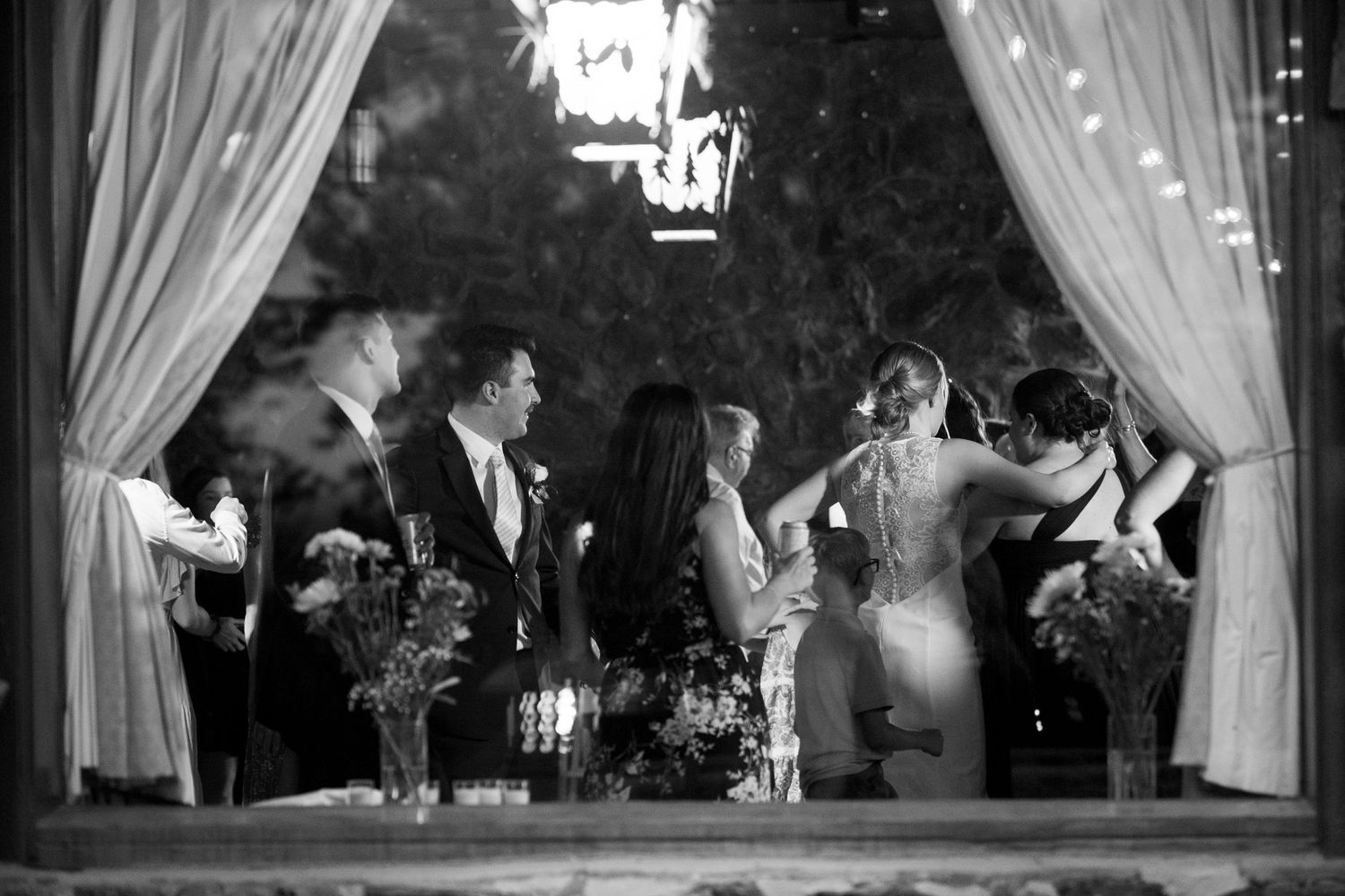 Black and white reception scene through draped curtains with chandelier and dancing guests.
