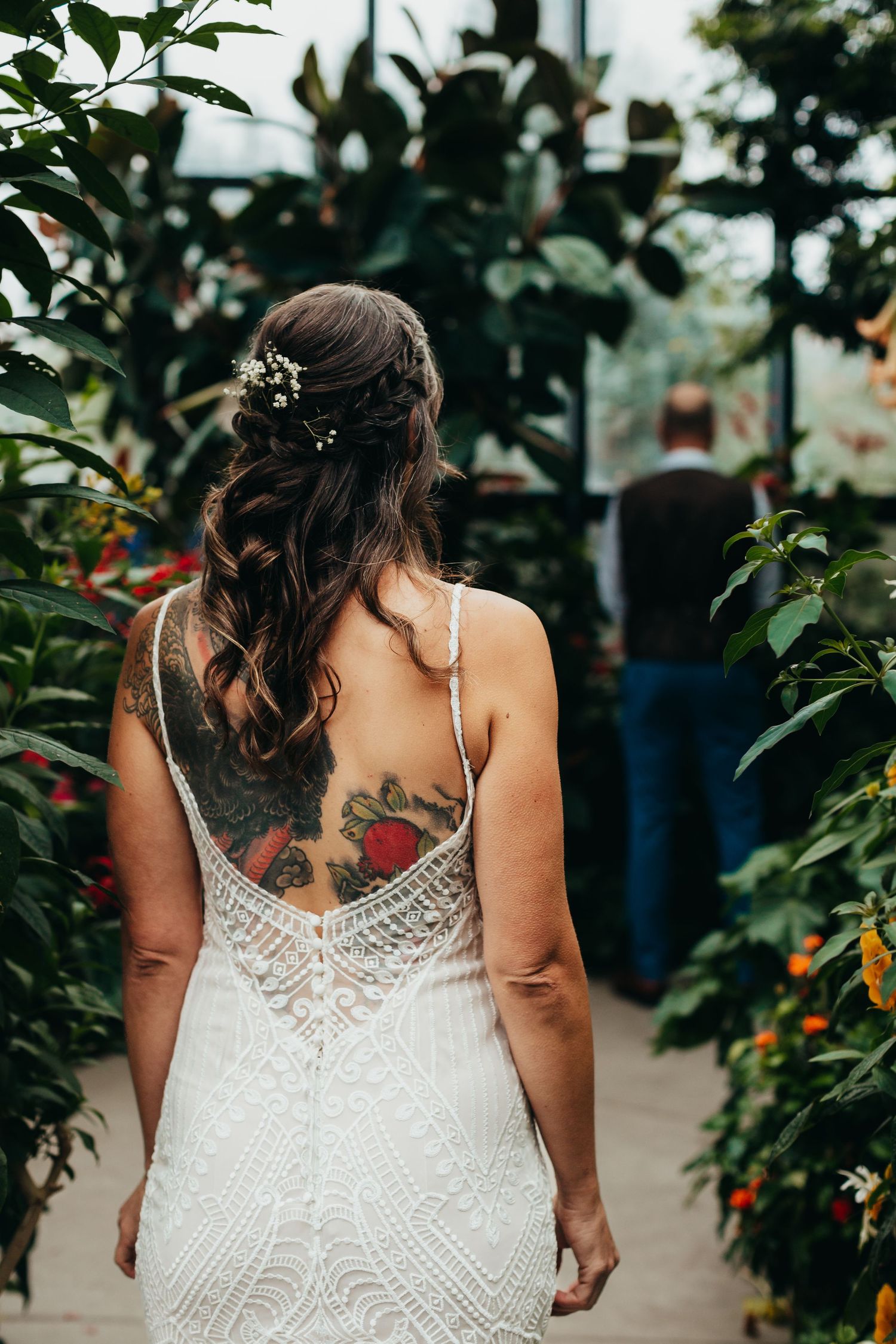 Back view of detailed lace wedding dress with visible tattoo in greenhouse setting.