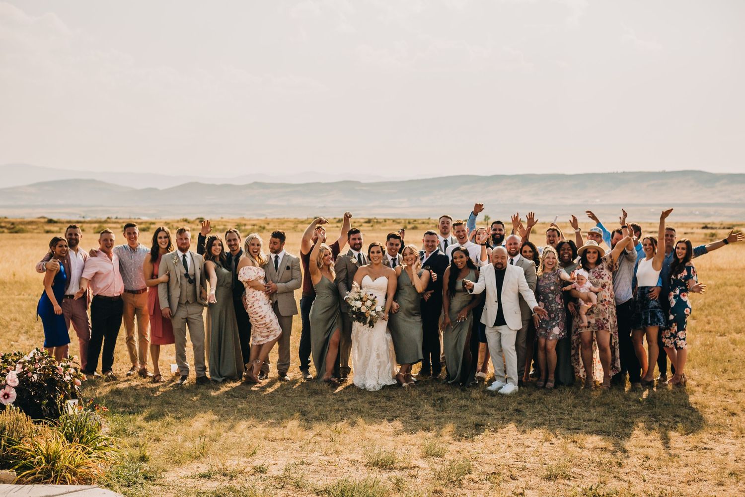 Large wedding party stands together in an outdoor field with mountains in the background during sunset.