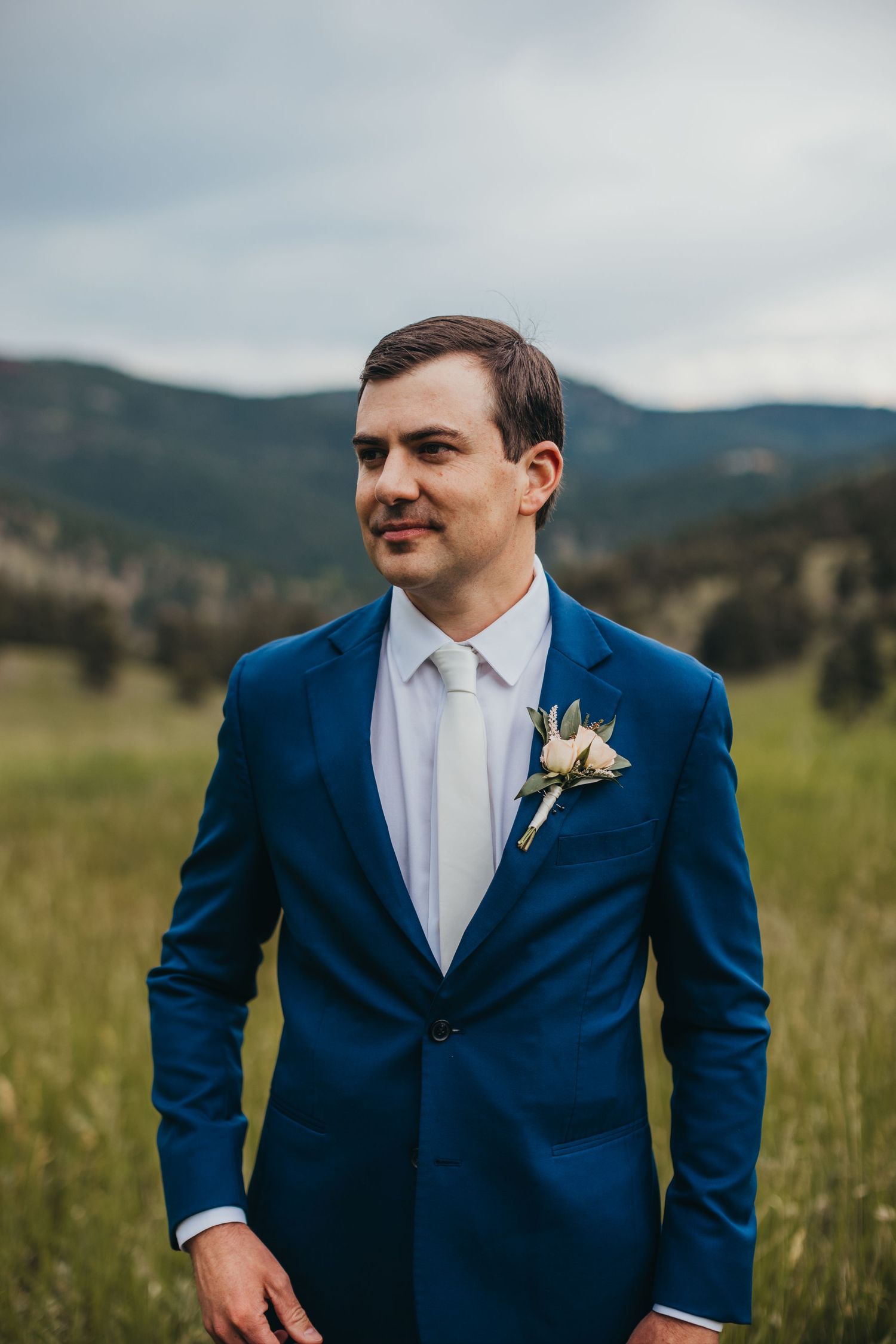 Groom in navy blue suit with white boutonniere against mountain landscape.