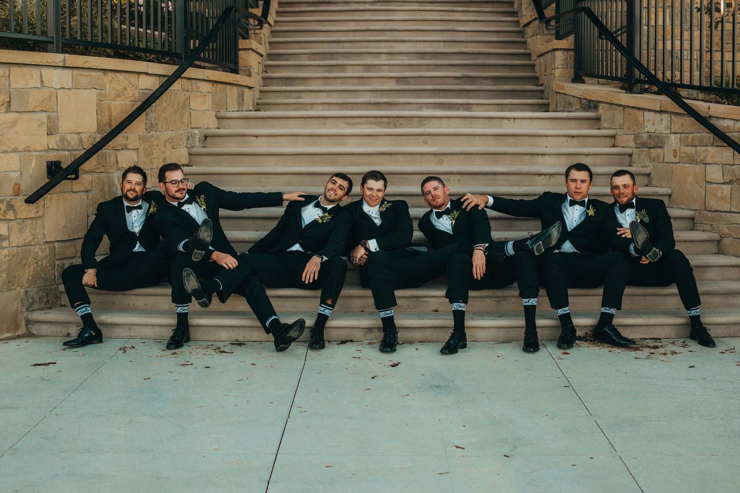 Groomsmen in black tuxedos sit casually on stone steps for a fun wedding party photo.