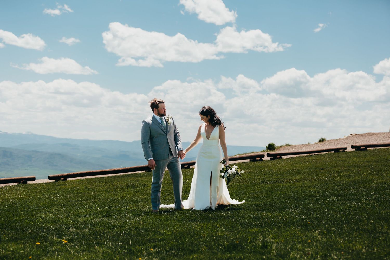 Newlyweds holding hands on hilltop with mountain vista behind them.