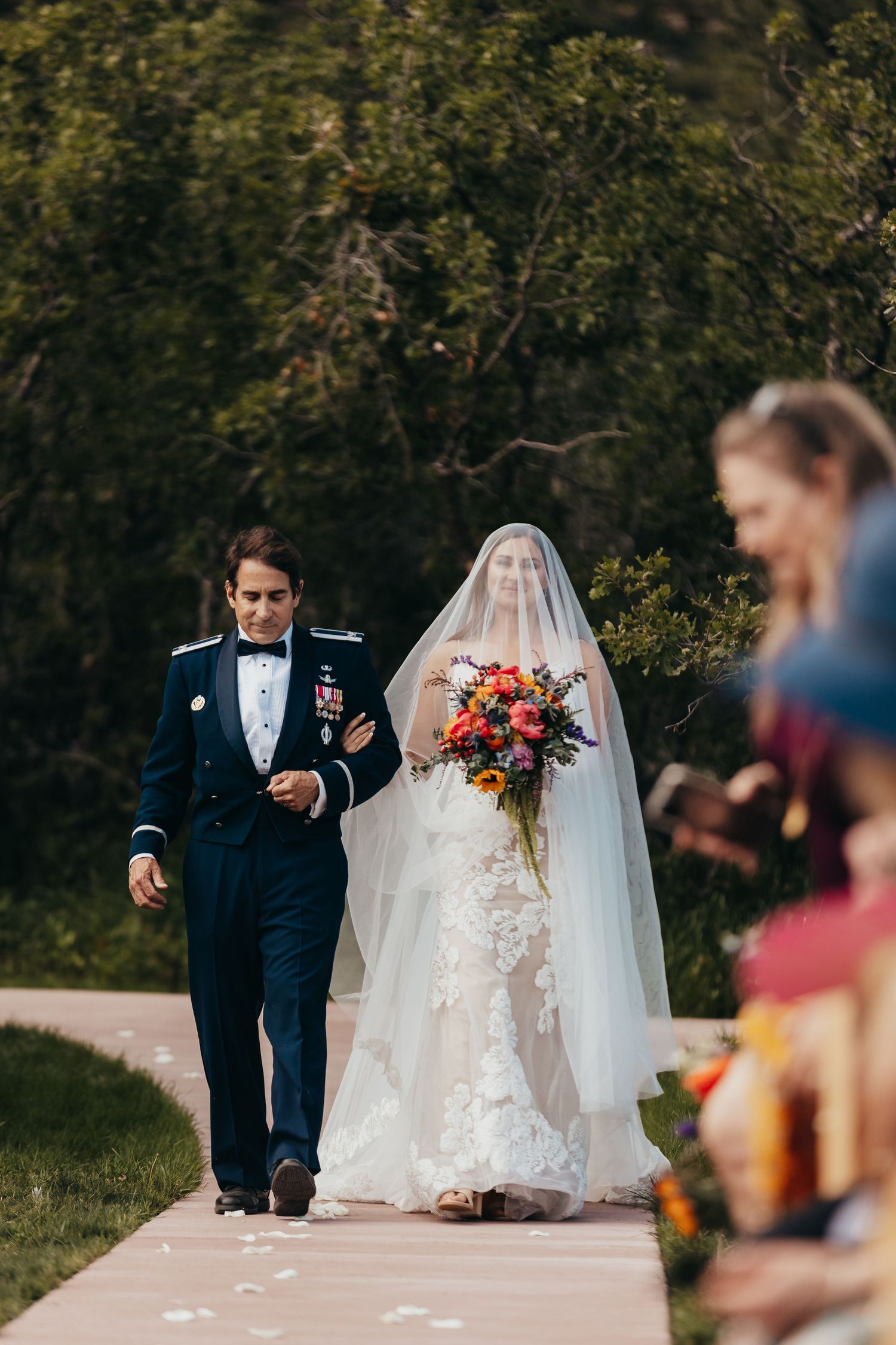 A wedding processional walks down an outdoor path with colorful bouquet and veil flowing.
