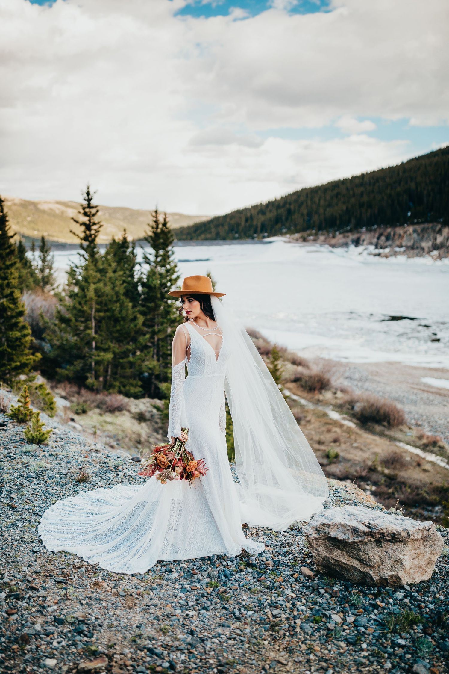 Bride in long-sleeve wedding dress and hat stands overlooking frozen mountain lake.