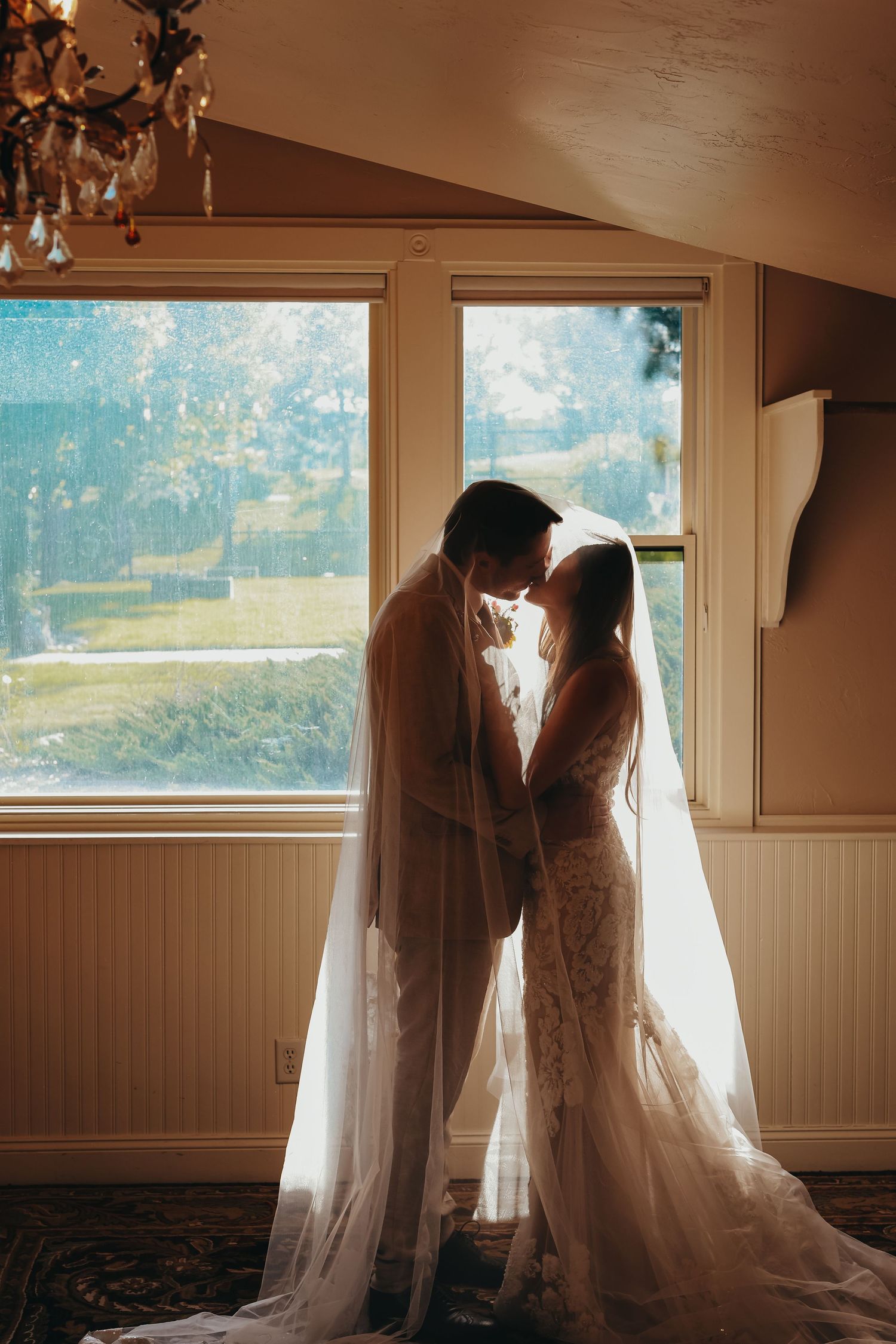 Silhouetted wedding couple embraces by window in romantic backlit scene.
