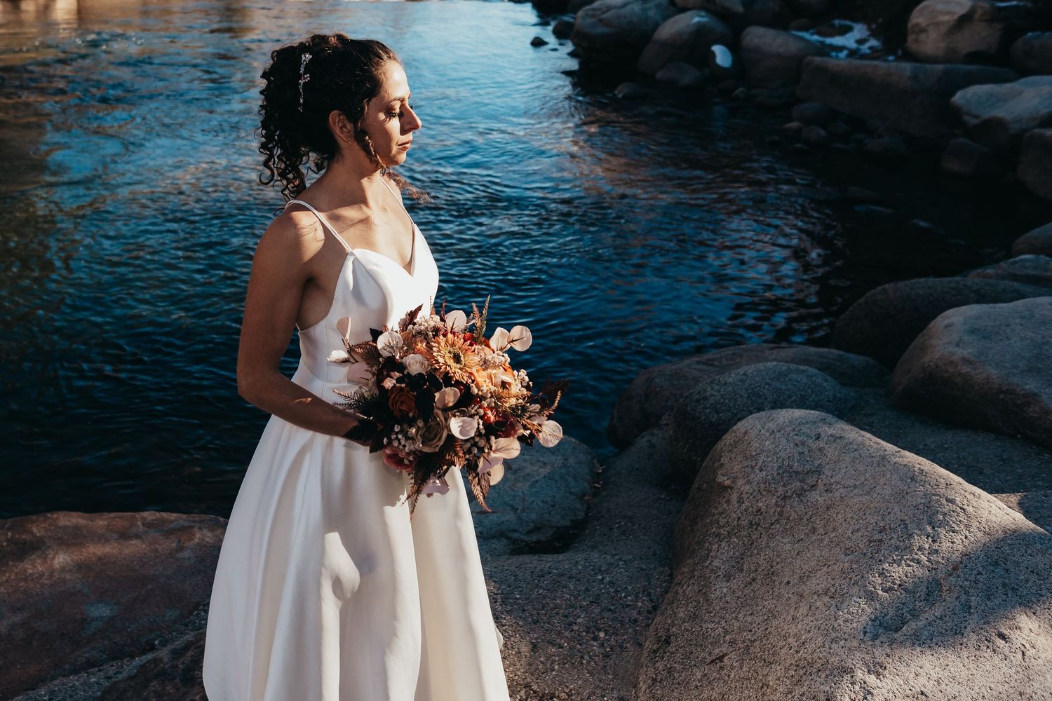 Bride in elegant white dress holding a peach-colored bouquet by a rocky riverside.