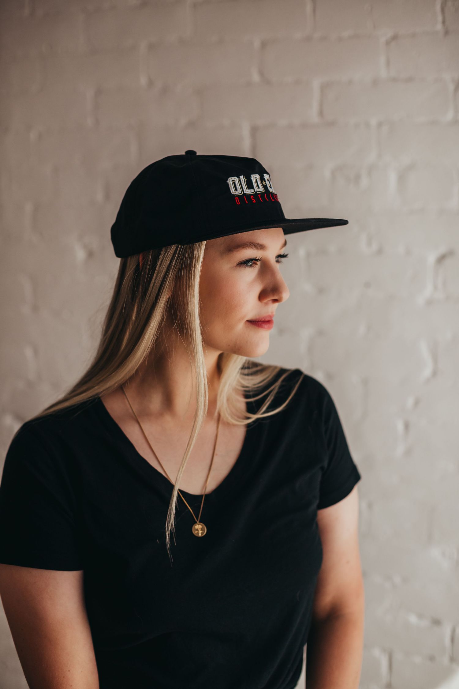 A thoughtful profile shot of someone wearing a black baseball cap and t-shirt with a delicate necklace.