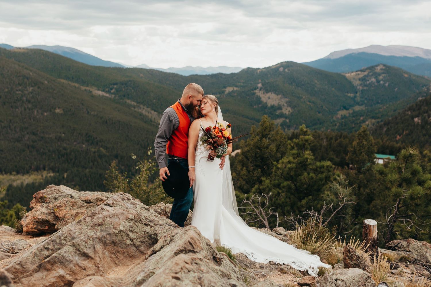 A couple embraces on a mountain overlook with dramatic mountain vistas in the background.