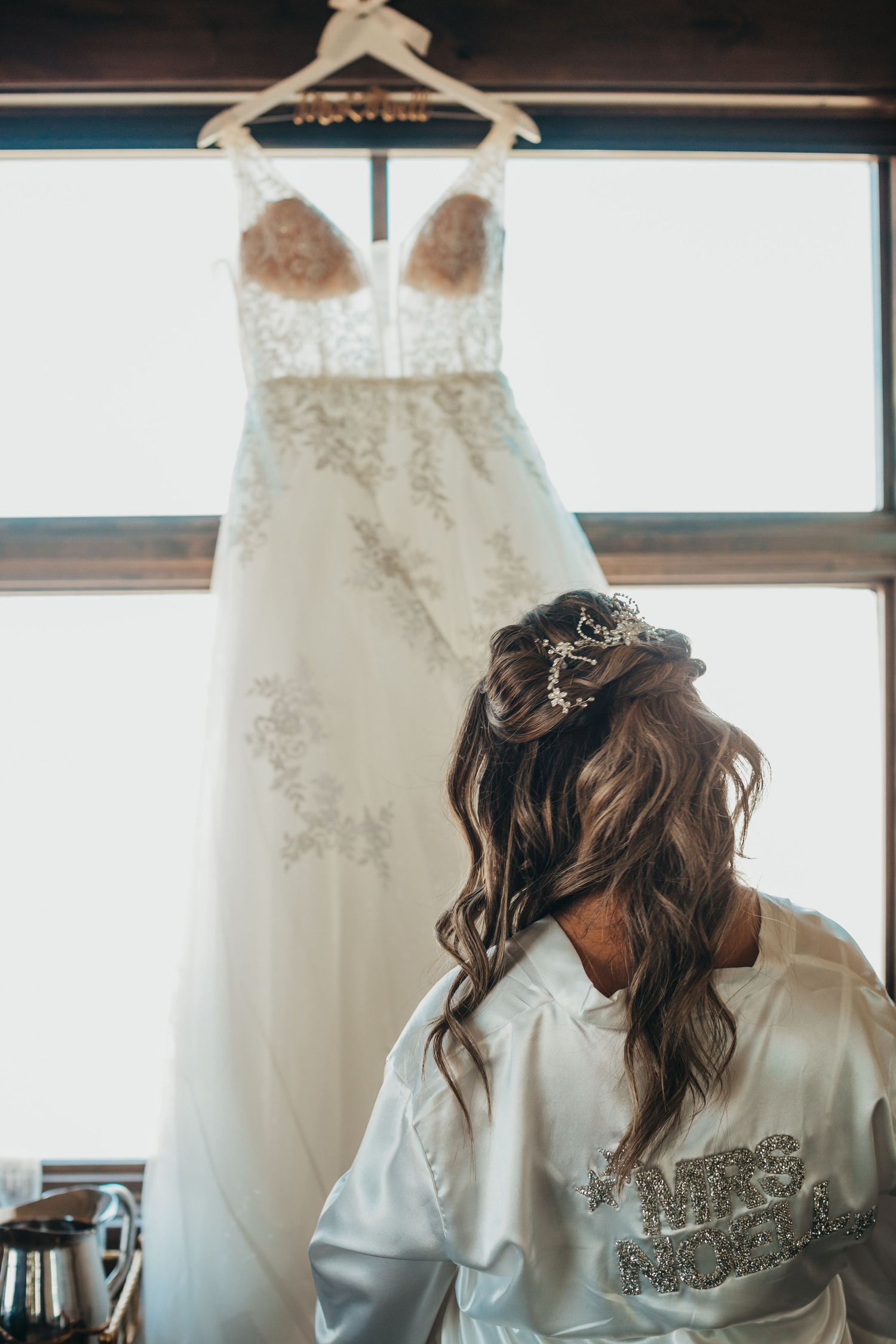 A white lace wedding dress hangs above someone in a bridal robe with styled hair.