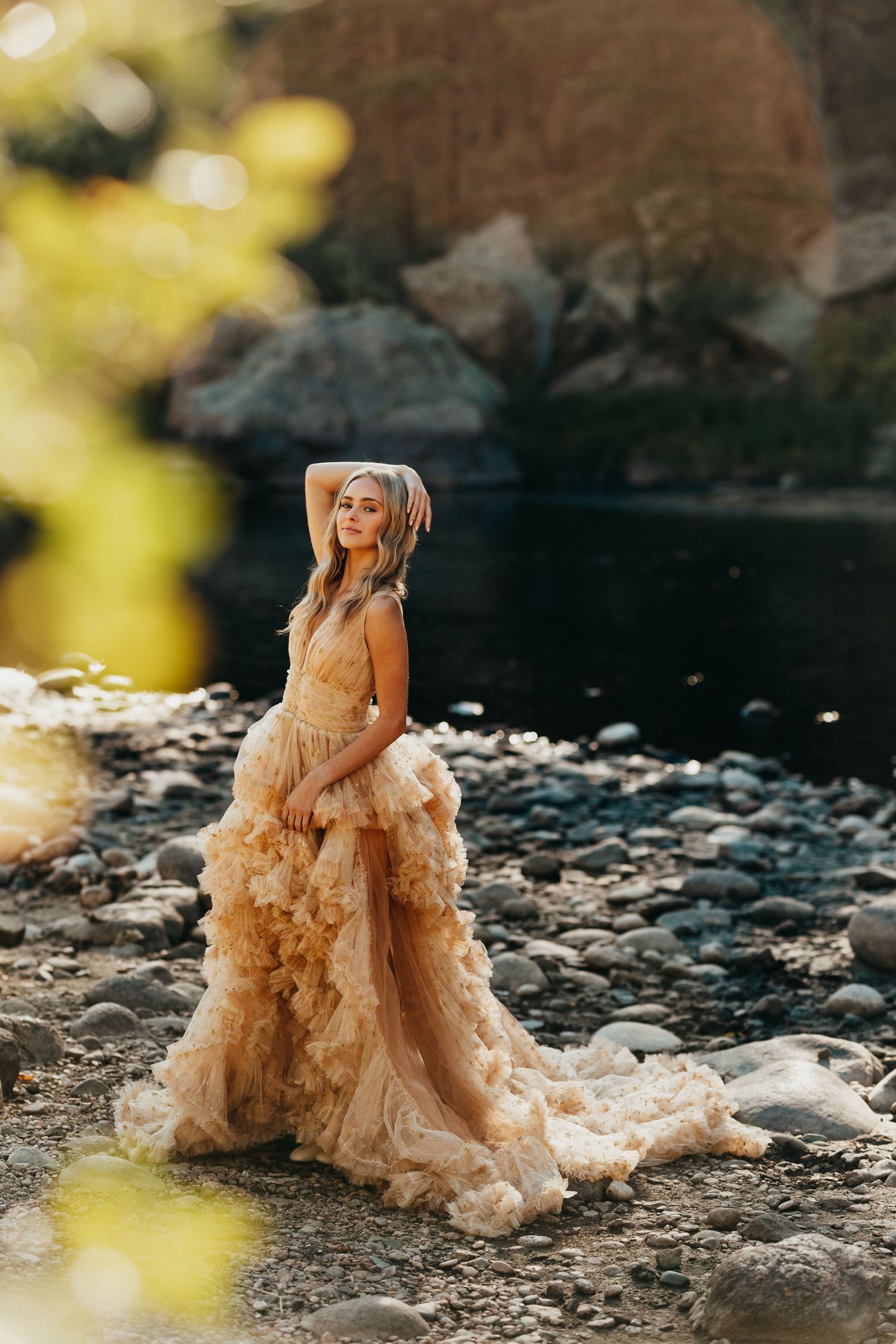 Someone poses in a flowing champagne-colored gown by a rocky riverbed.