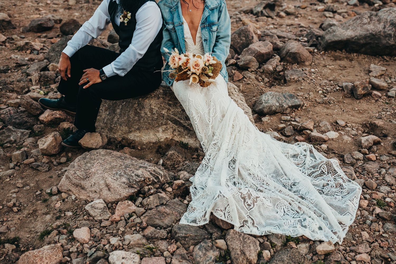 Closeup of elegant lace wedding dress train and bouquet against rocky terrain at mountain lakeside.