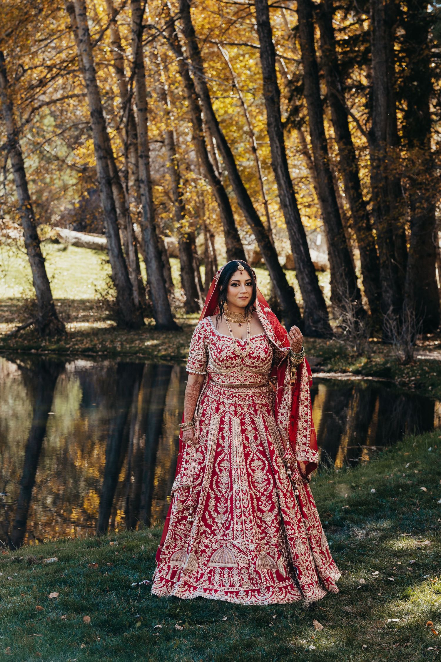 Traditional red bridal lehenga with intricate embroidery photographed by a reflective pond in autumn forest setting.
