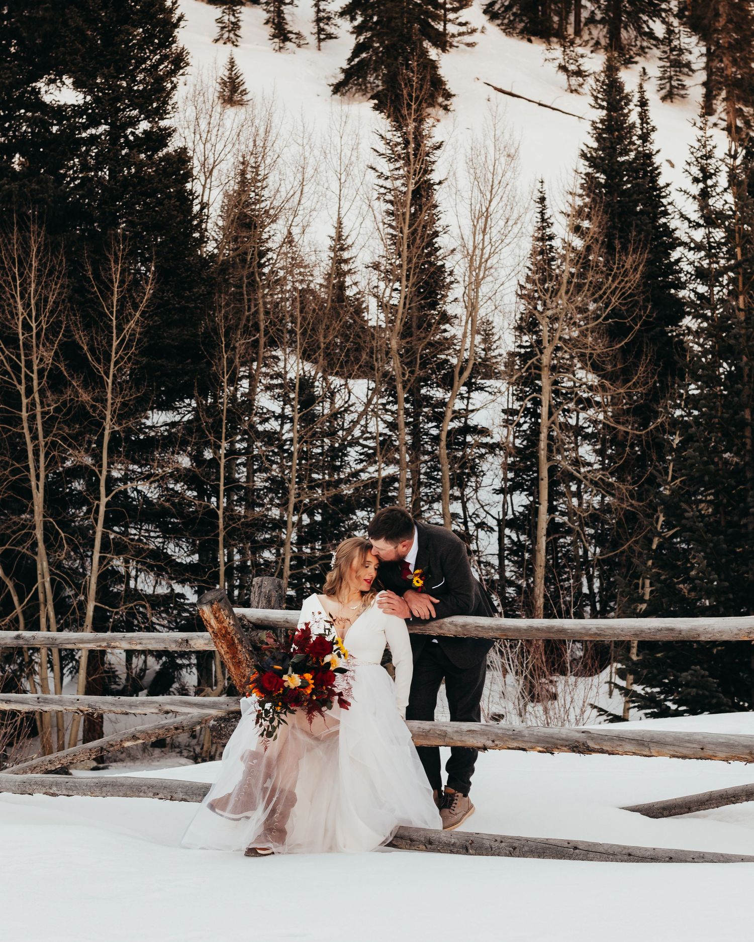 A couple shares a romantic moment on a snow-covered deck surrounded by evergreen trees.