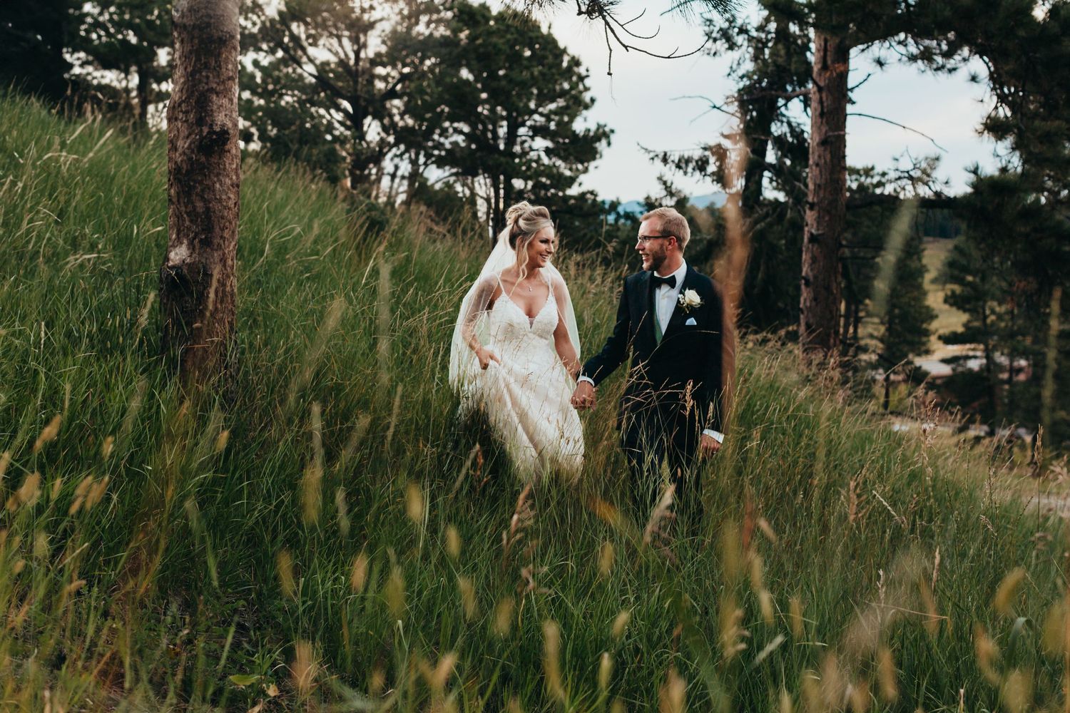 Couple walking through tall grass on hillside with pine trees in background.