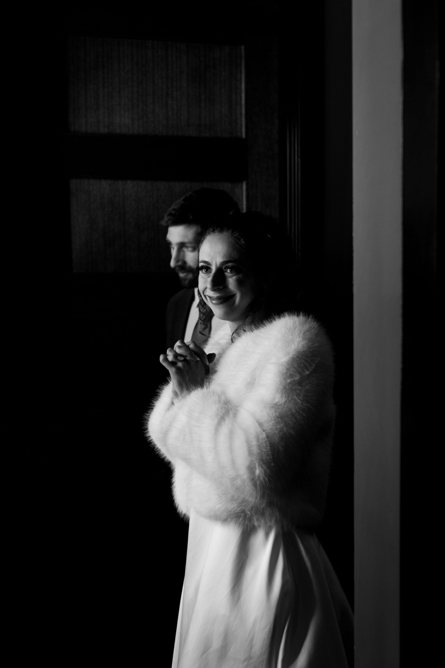 A romantic black and white portrait of newlyweds by a window in winter attire.