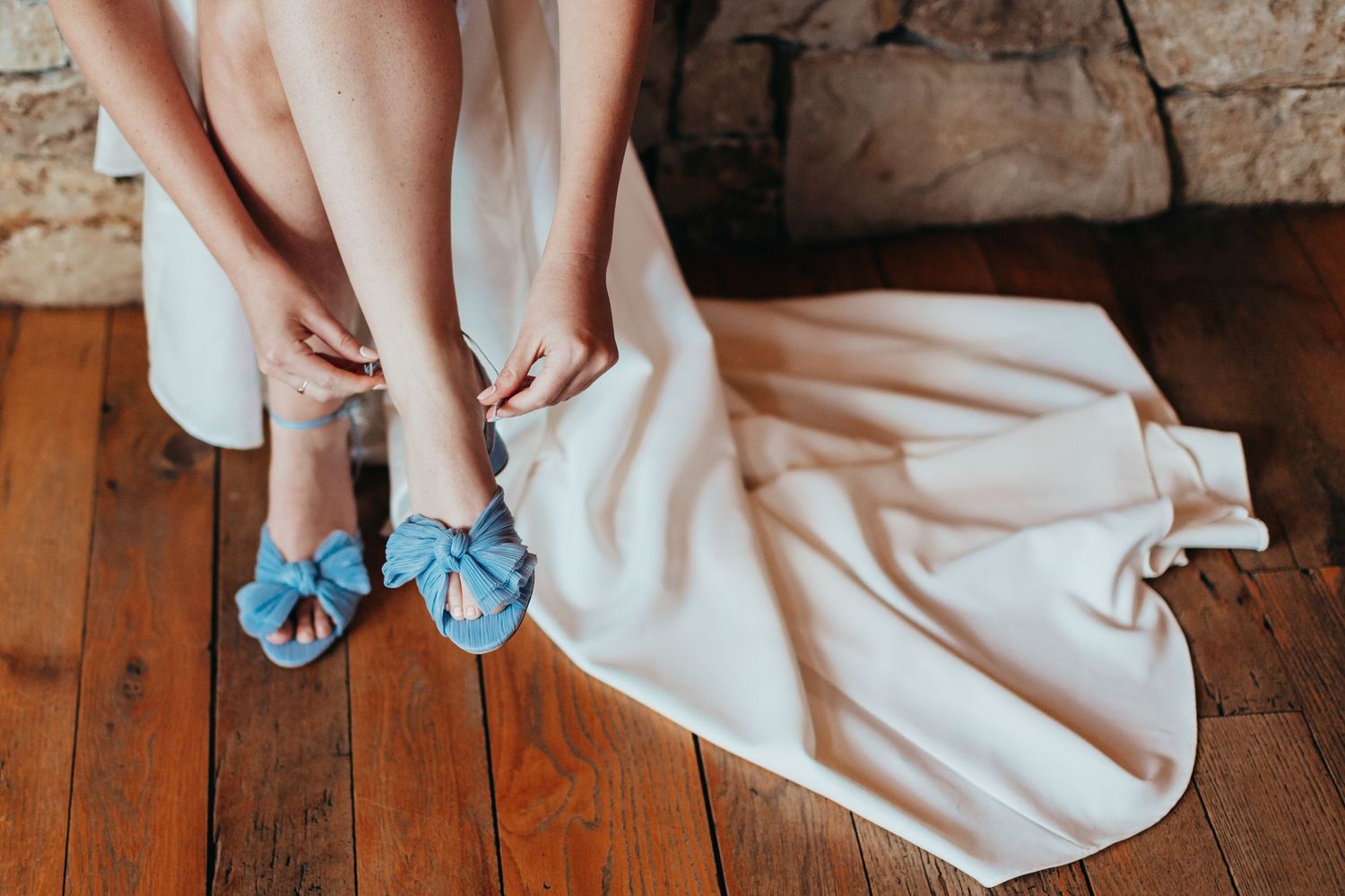 Blue shoes with bows peeking out from under a flowing white dress on wooden floor.