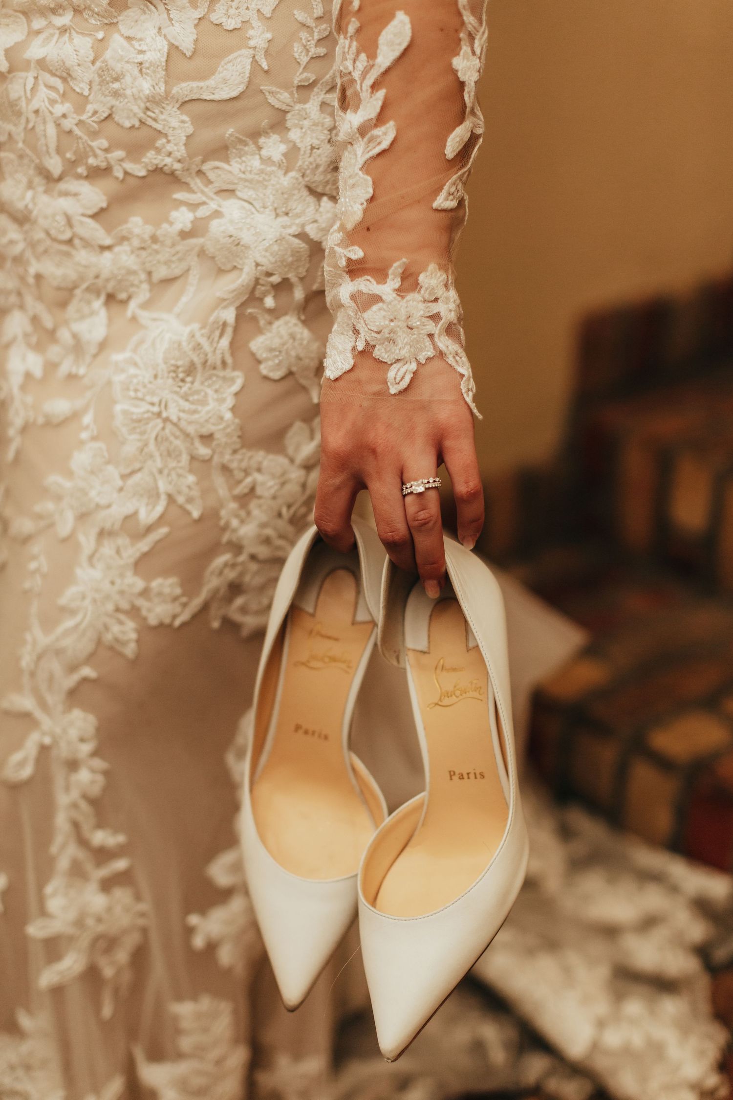 Elegant white bridal shoes held by hand wearing lace sleeve dress and engagement ring.