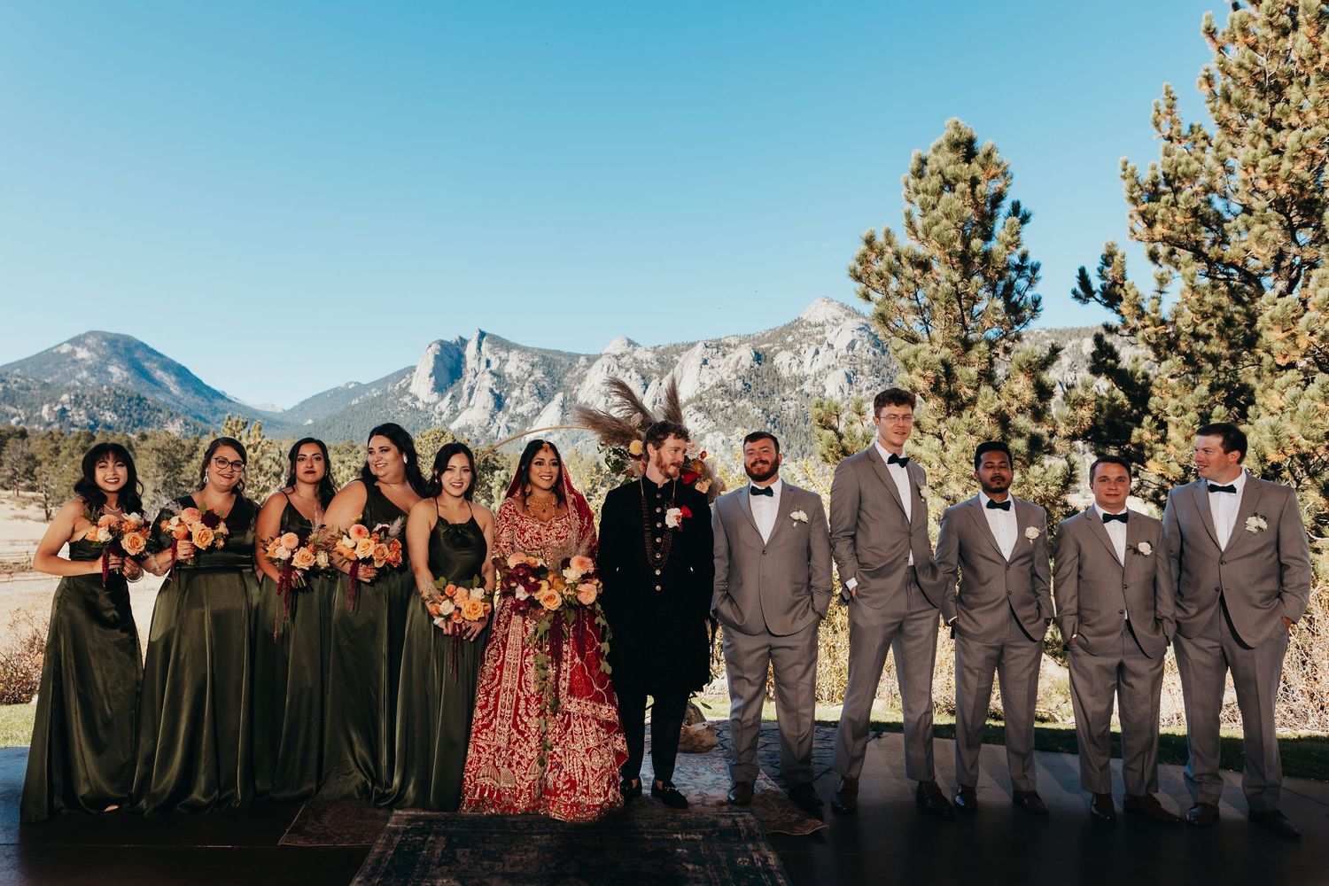 A wedding party in sage green and gray attire poses with mountain scenery in the background.
