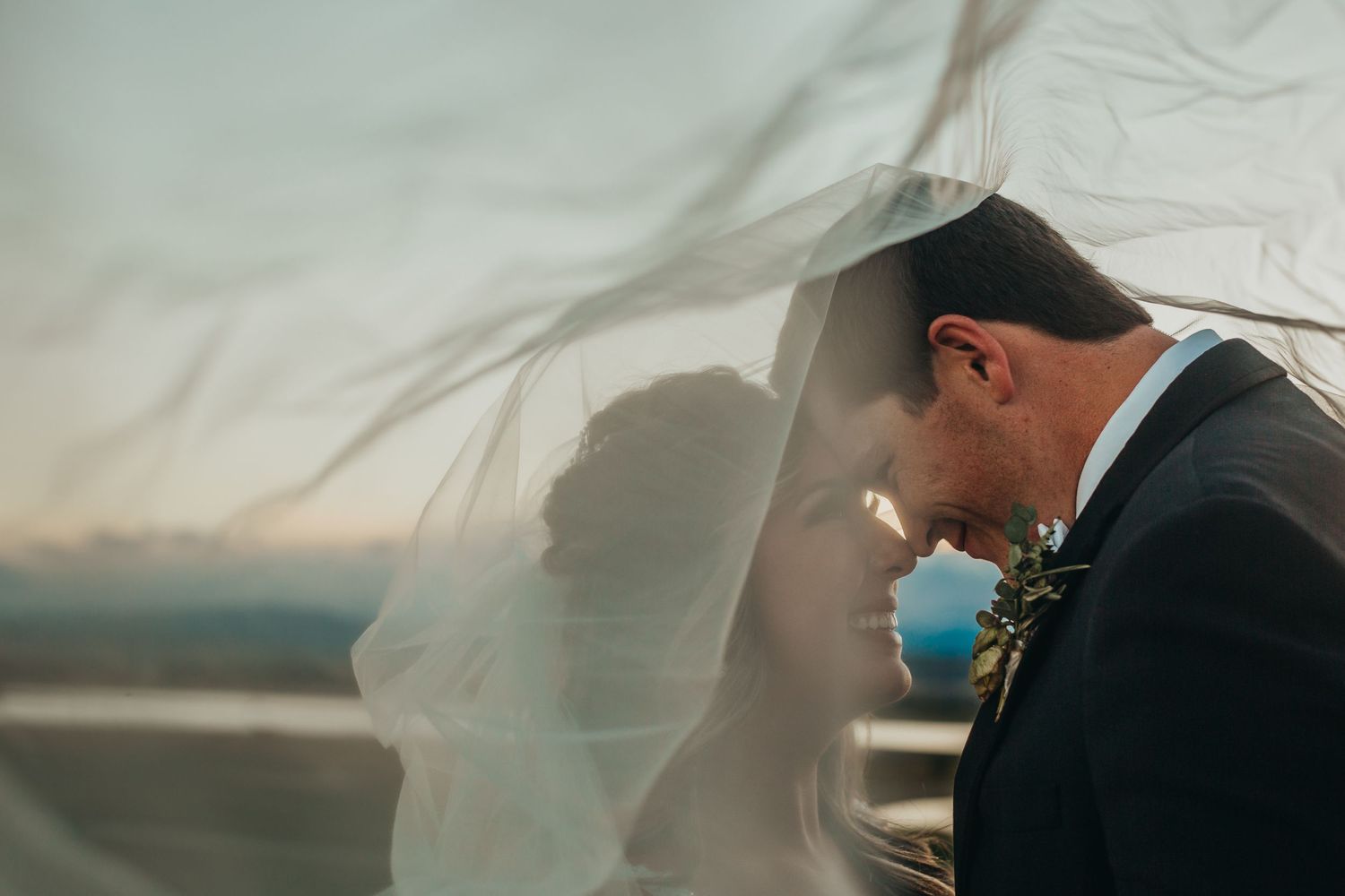 A tender moment between newlyweds as wind catches the bride's veil during sunset.