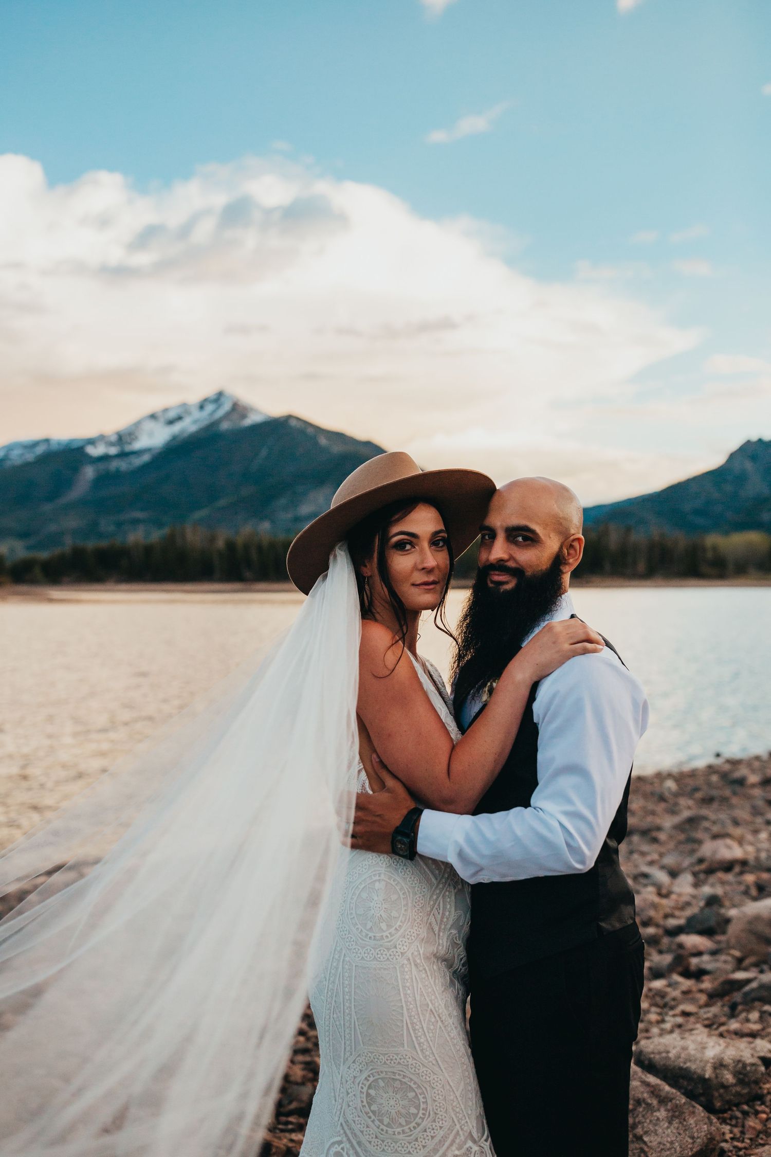 Mountain lake wedding portrait with brown hat and flowing veil against scenic backdrop.