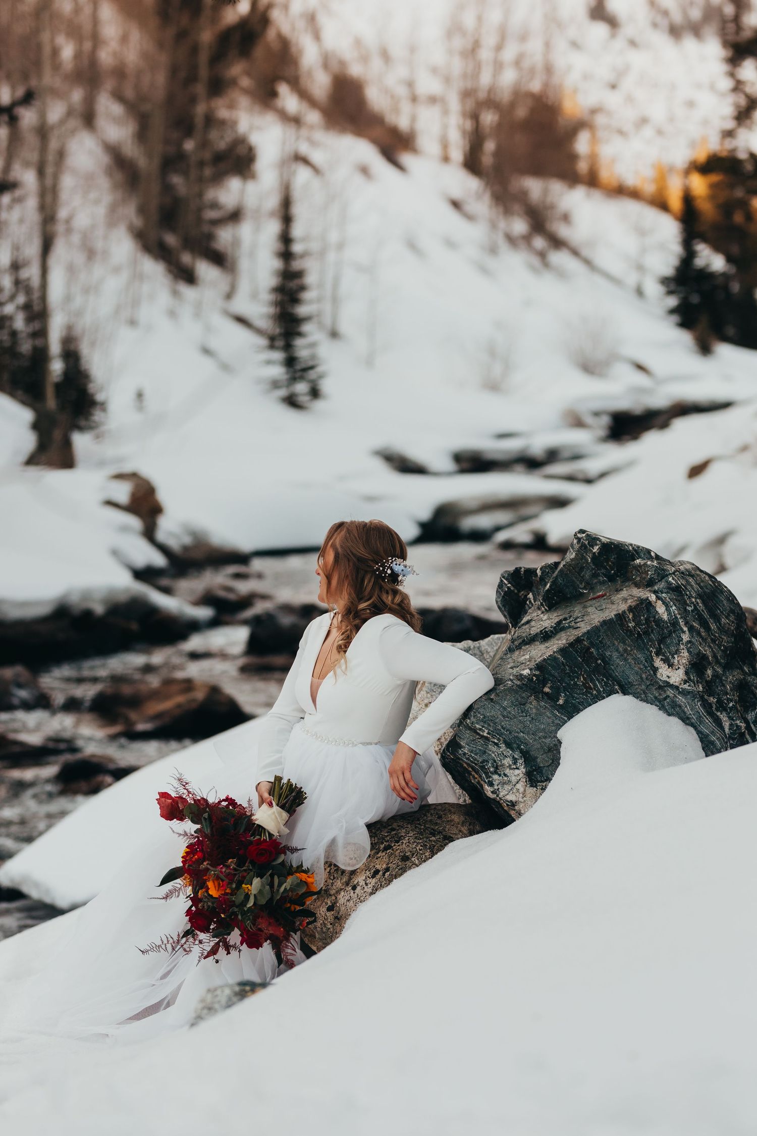 Bride in white gown sits on snowy rocks with colorful bouquet near mountain stream.