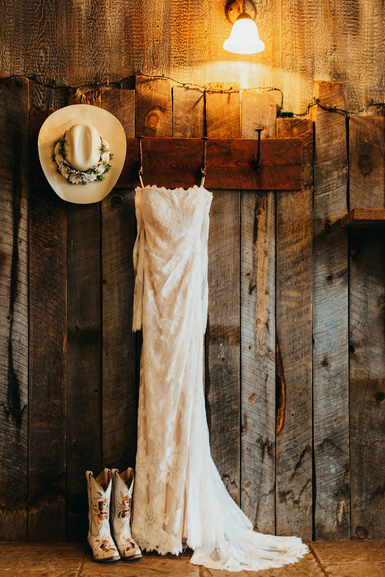 White wedding dress, cowboy boots and floral hat displayed against rustic wooden wall.