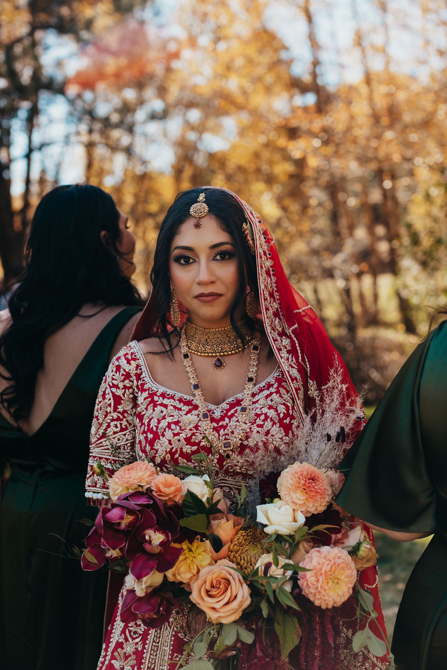 Bride in traditional Indian attire holds bouquet surrounded by autumn foliage.